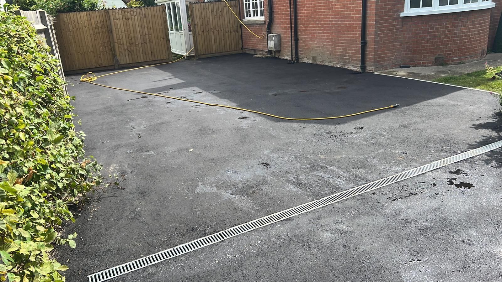 Black asphalt driveway with yellow and black curbing, beside brick house and hedge.