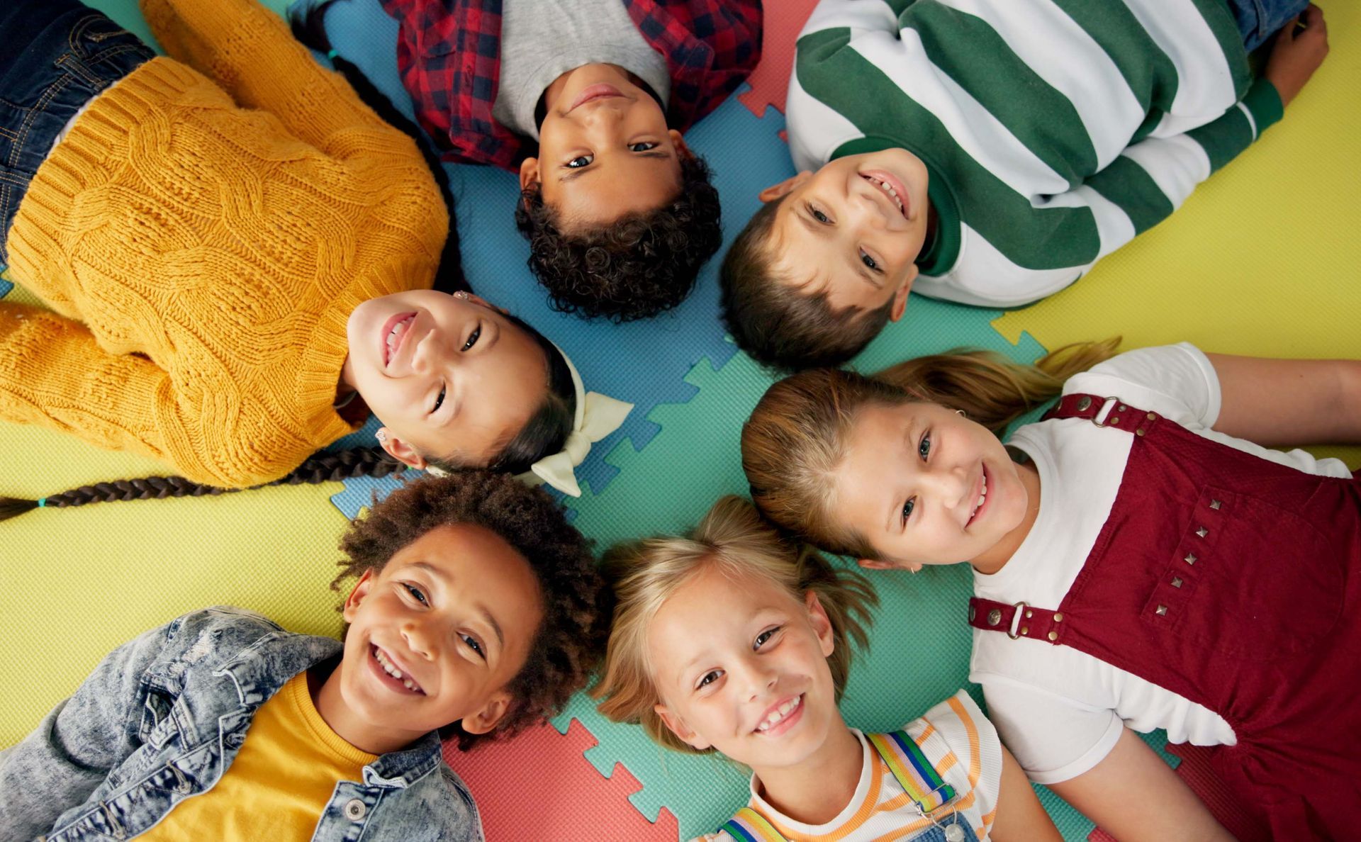 Group of children lying in a circle, smiling. On a colorful playmat.