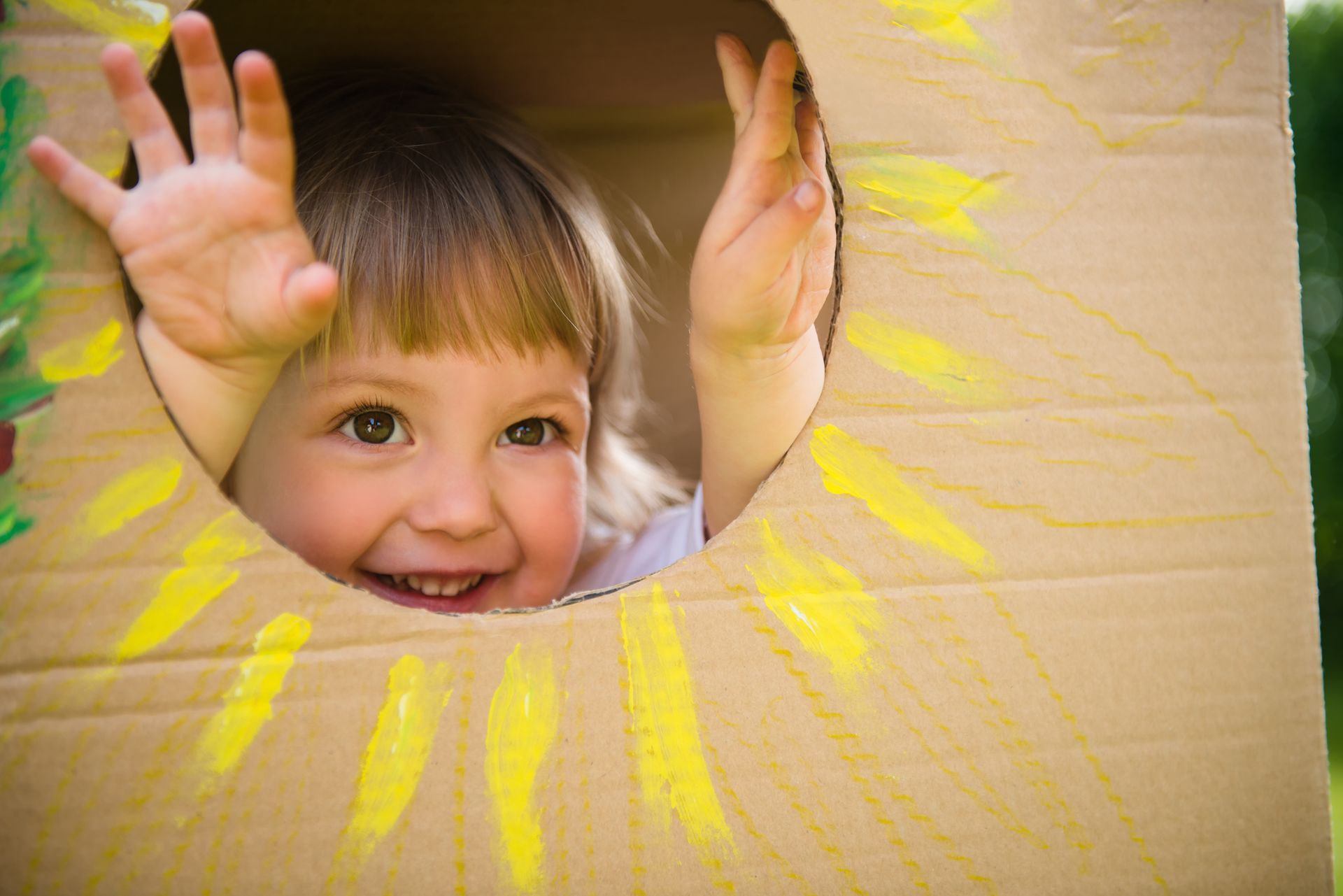 Young child with a smiling face, peeking out of a cardboard box with a painted sun.