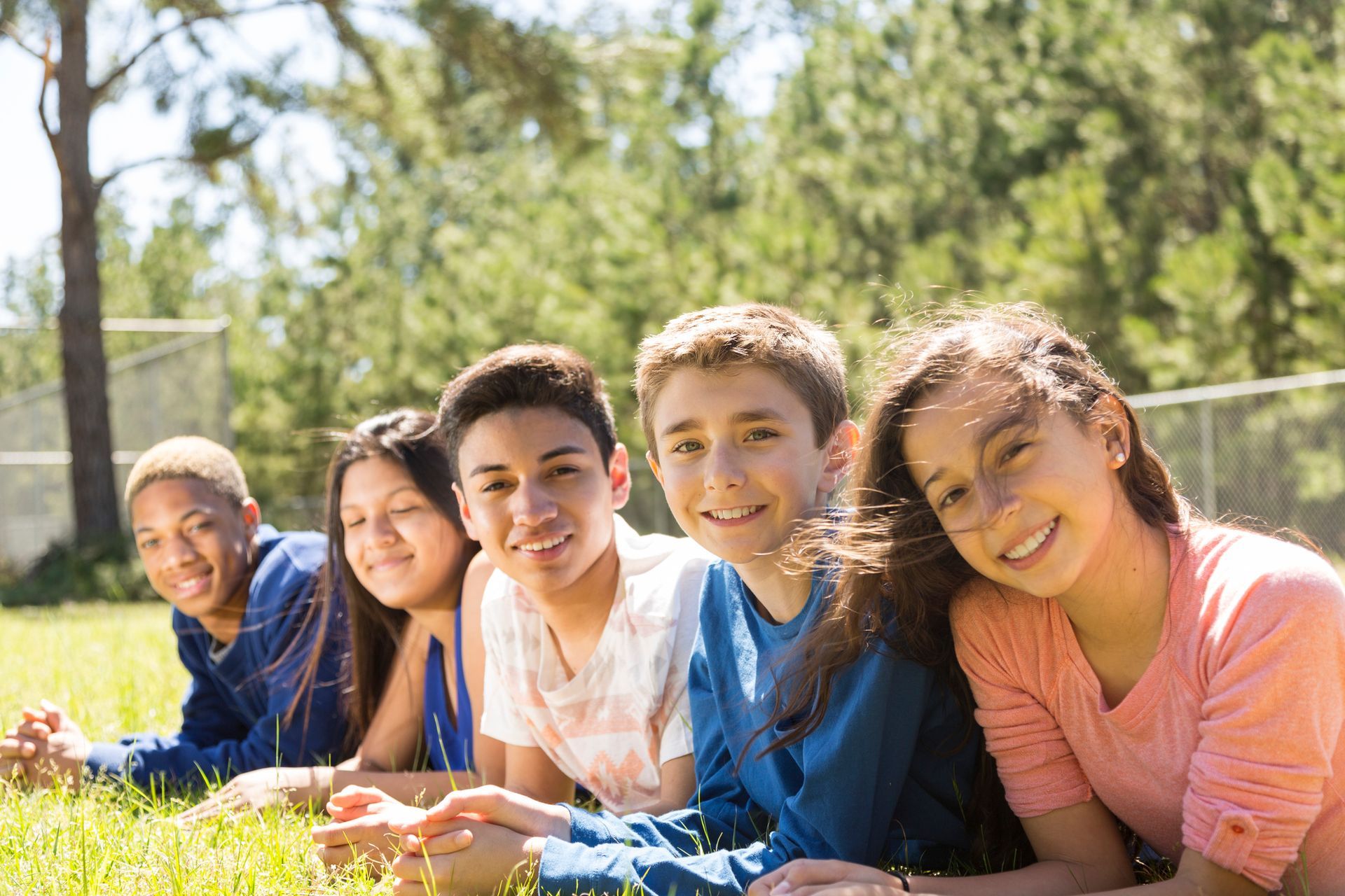 Five diverse teens smiling while lying on grass outdoors.