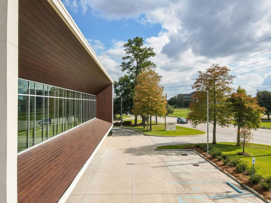 Modern building with brick wall and glass windows beside a paved walkway and trees under a cloudy sky