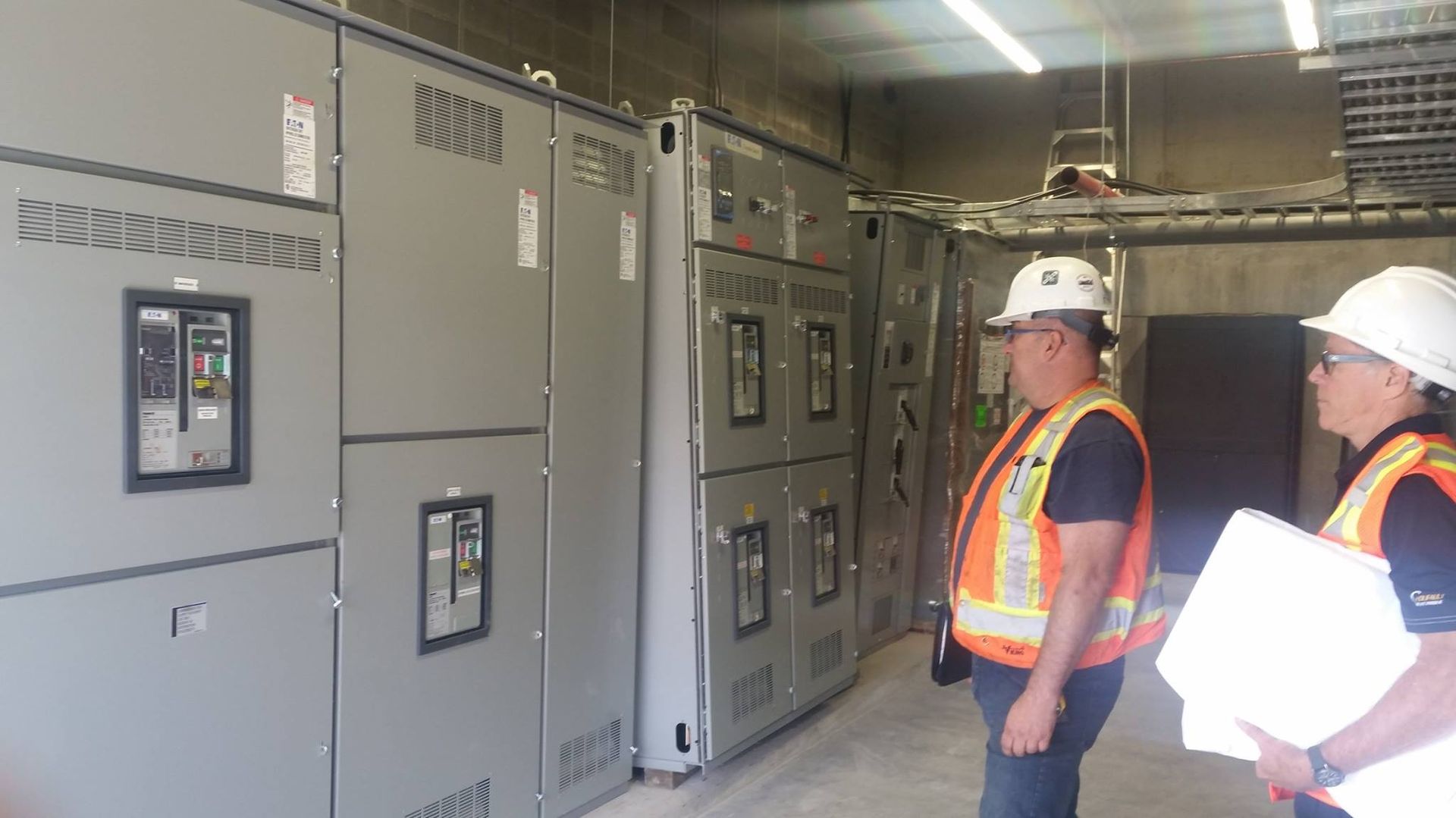 Deux hommes portant des casques de chantier se tiennent dans une pièce avec des boîtiers électriques.