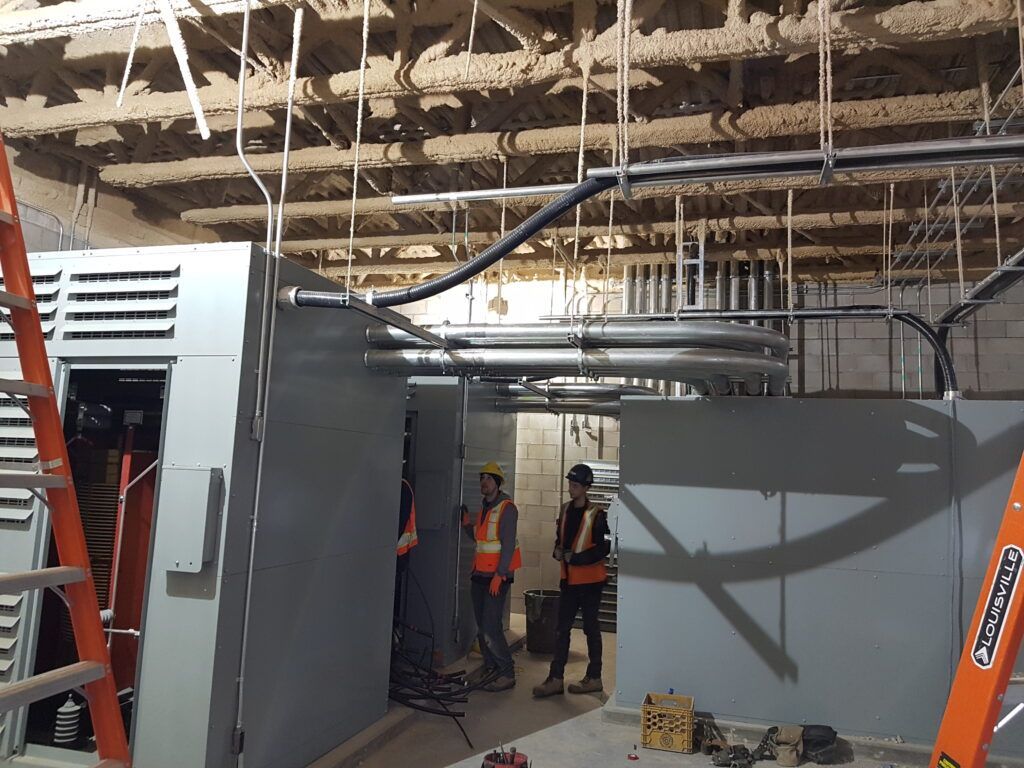 A group of construction workers are standing in a room with pipes hanging from the ceiling.
