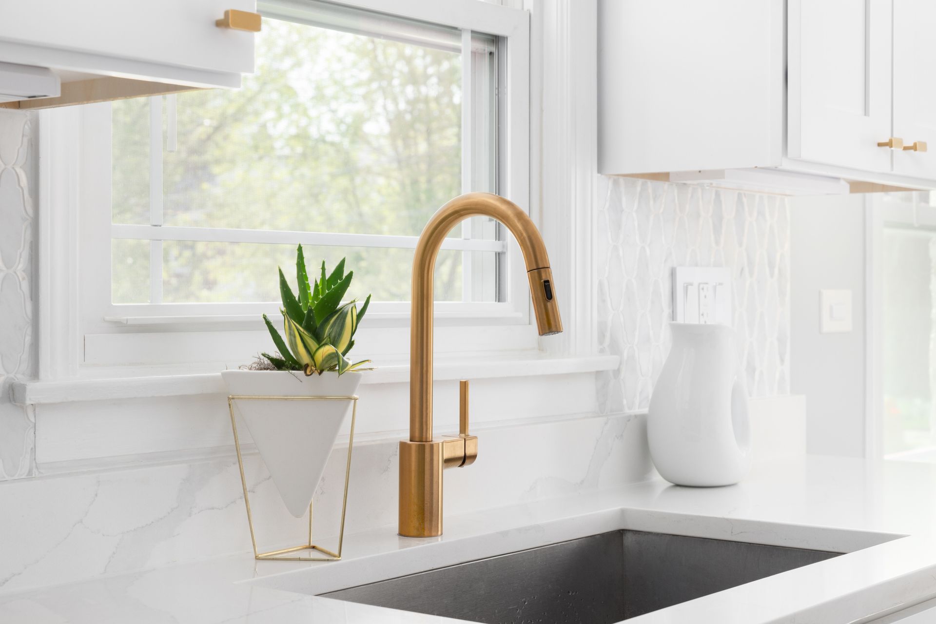 Bright white kitchen sink with brass faucet, potted plant, and vase by a sunlit window