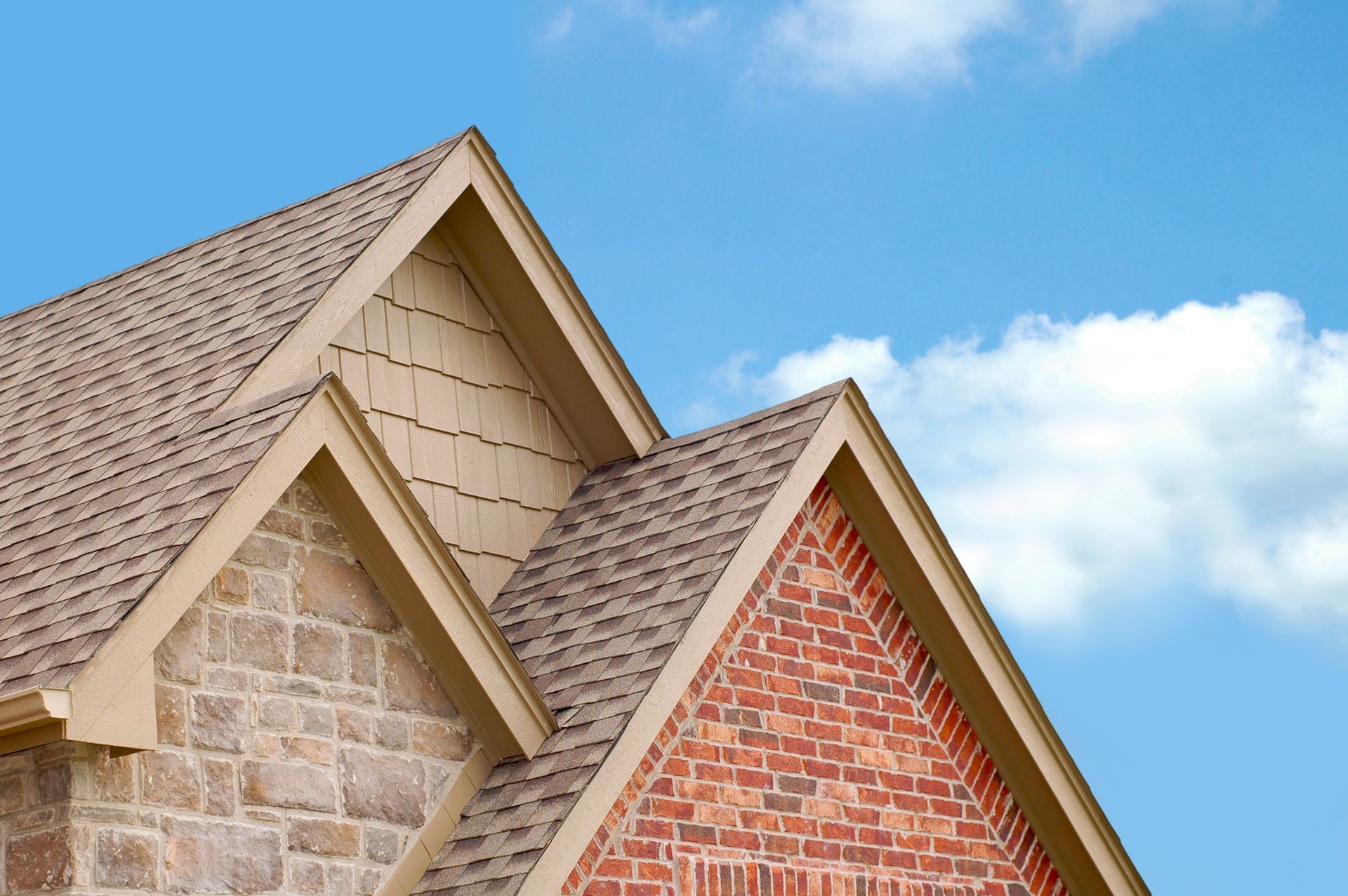 Two house roofs with beige shingles and stone and red brick walls under a blue sky.