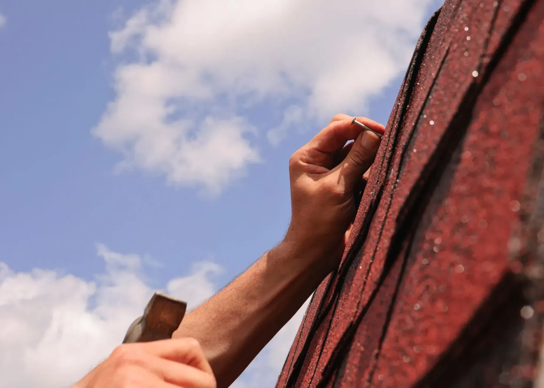 Hand applying shingles to a red roof against a blue sky with clouds