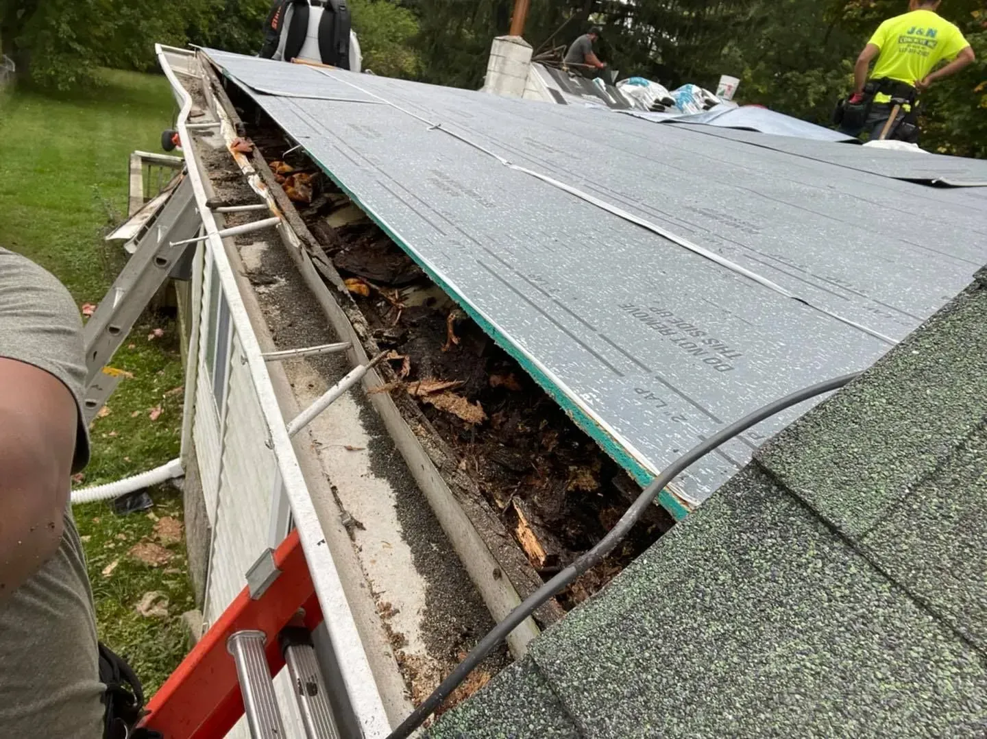 Workers replacing a roof, lifting gray shingles to reveal rotting wood along the edge