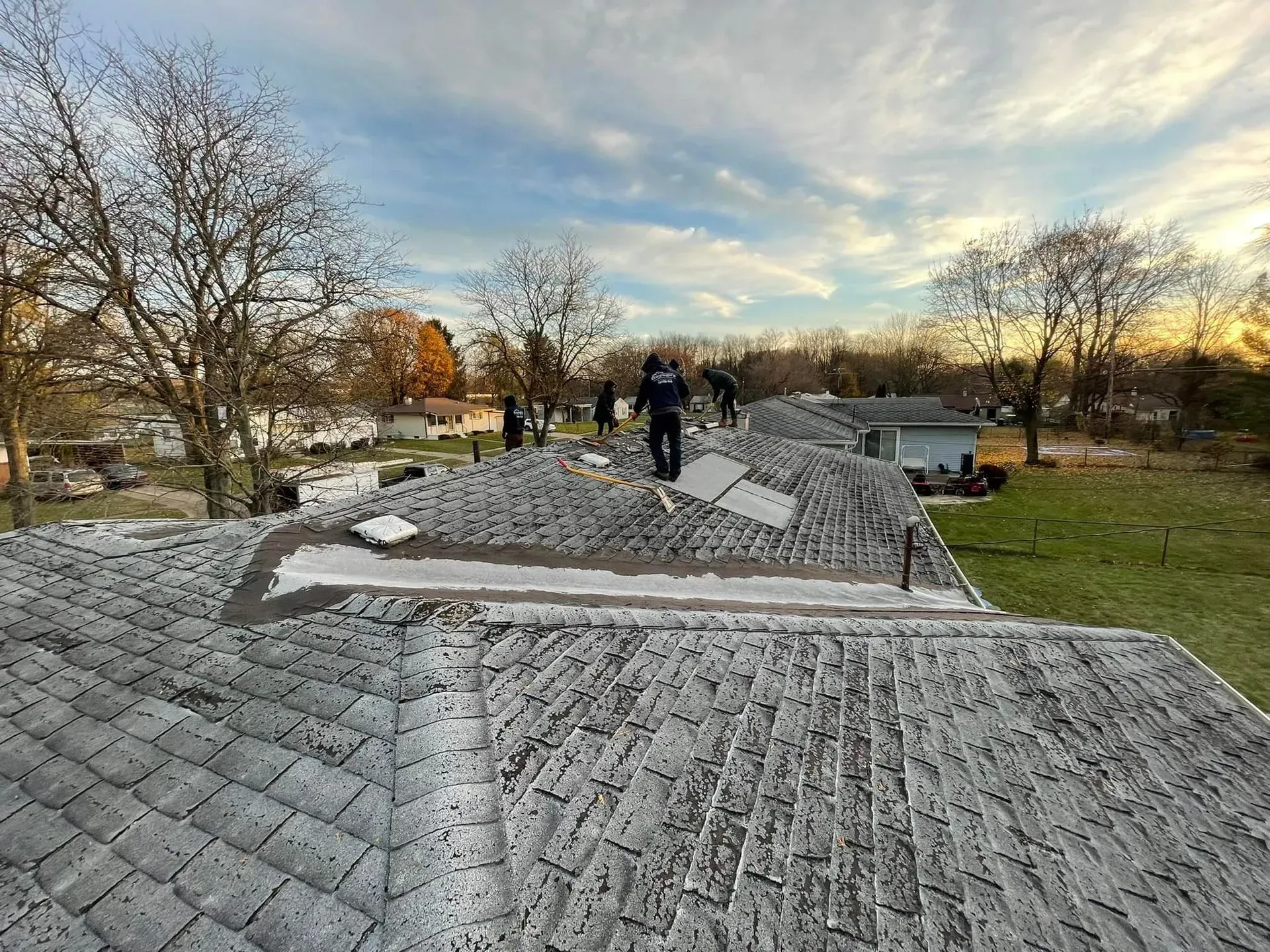 Workers installing gray roofing on a house under construction in a leafless yard at sunset