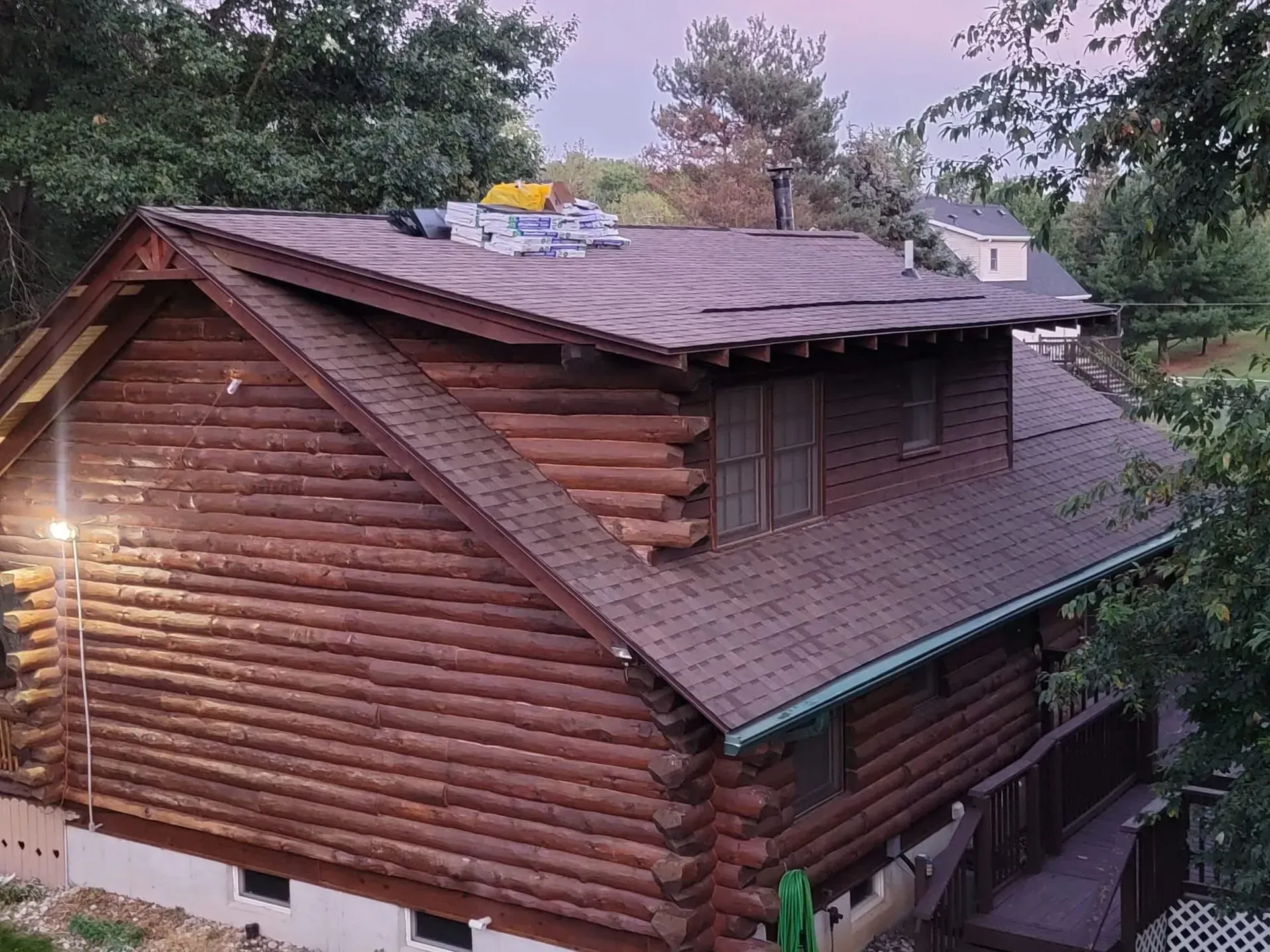 Log cabin with a shingled roof and rooftop equipment, surrounded by trees at dusk