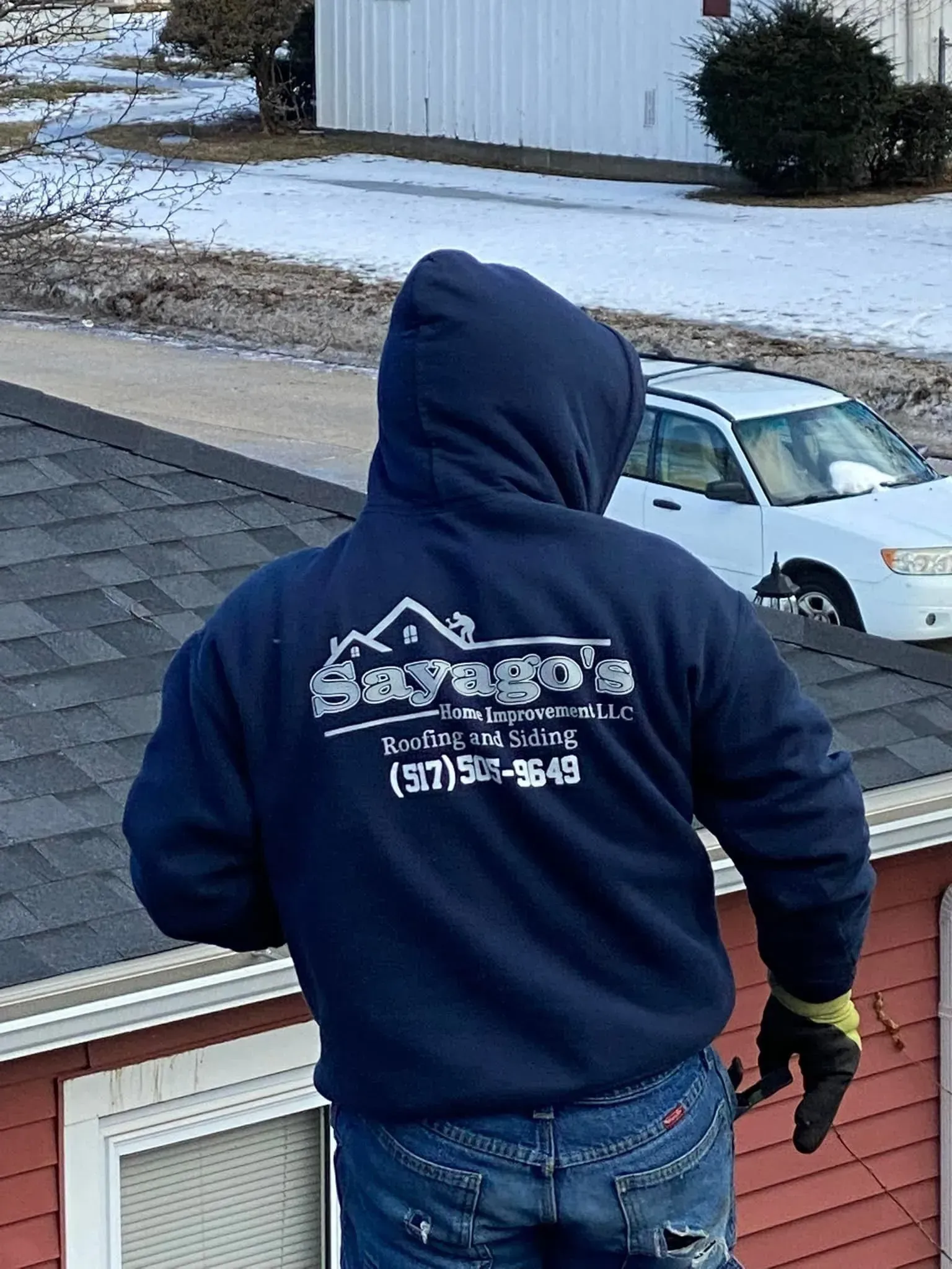 Person in a navy hoodie standing on a rooftop, looking down at a snowy driveway and parked car.