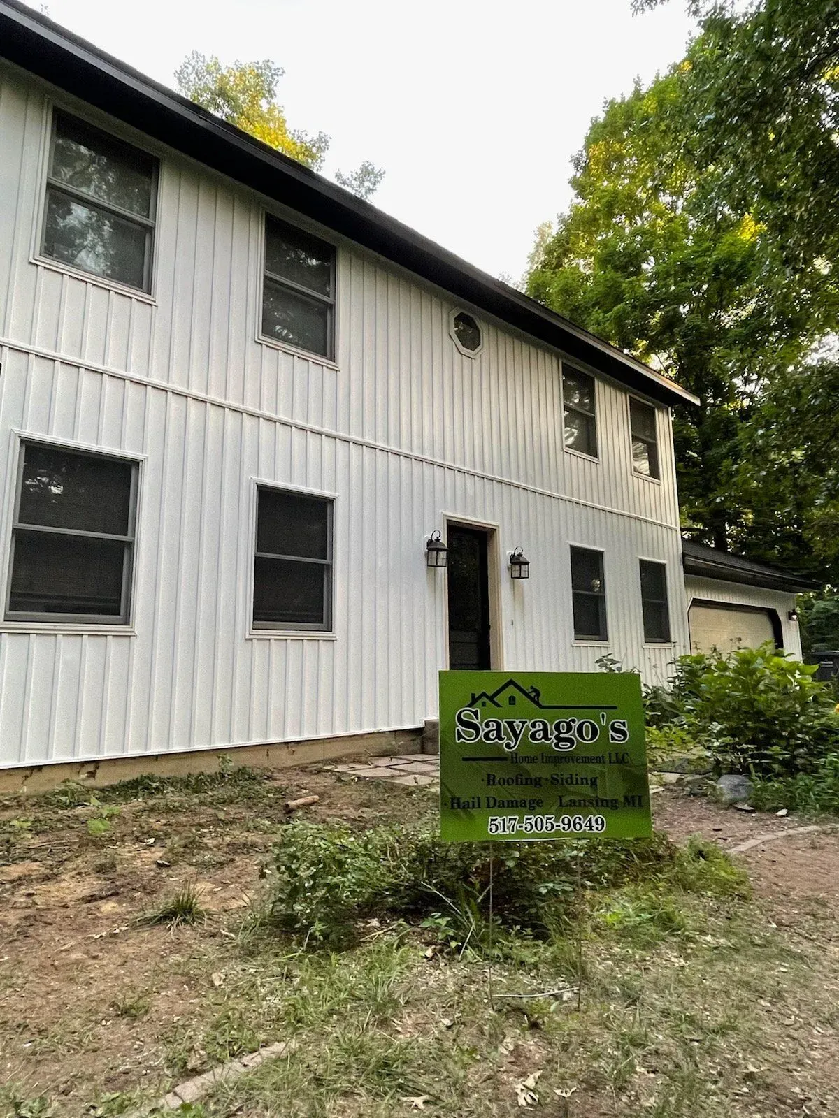 White two-story building with dark windows and a green sign in front, surrounded by trees.