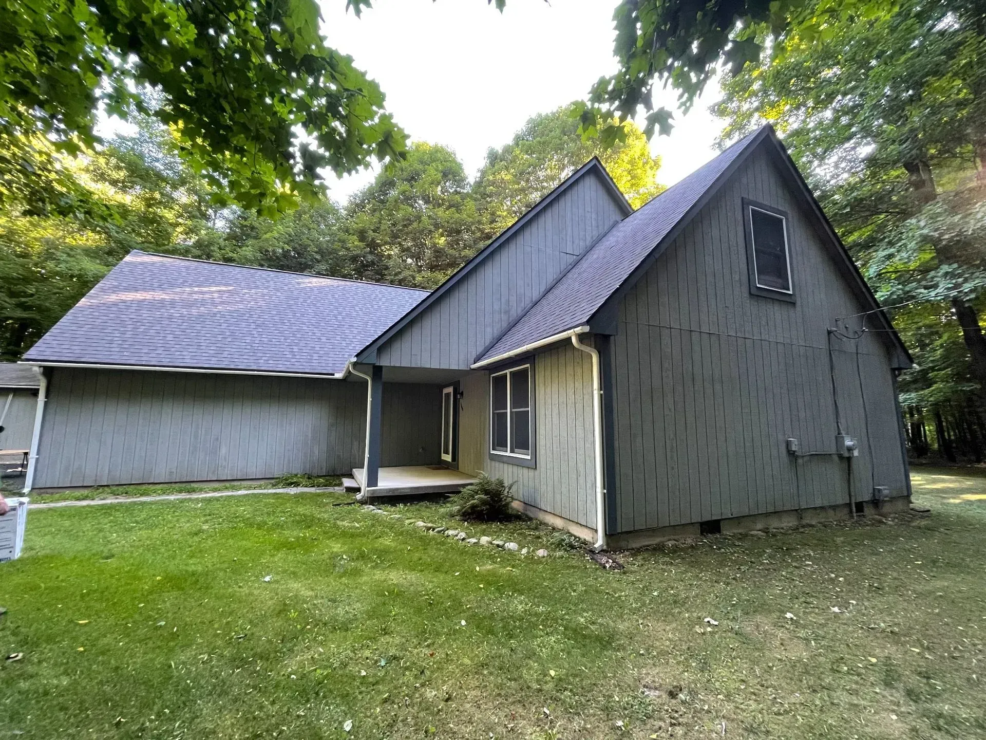 Gray house with steep roofs beside a larger barn-like building in a grassy yard surrounded by trees