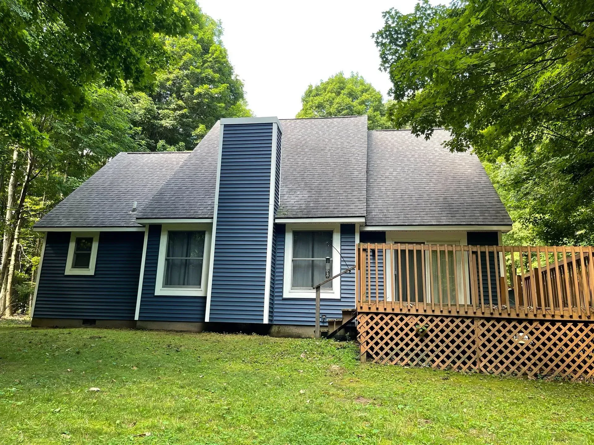 Blue house with a gray roof and wooden deck, surrounded by green trees and lawn