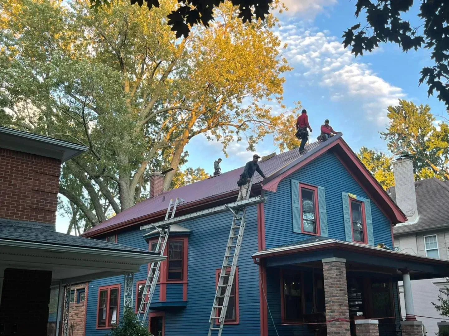 Workers on a blue house roof using ladders, surrounded by tall autumn trees and a partly cloudy sky