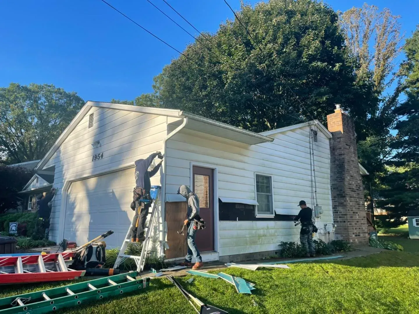 Workers on a ladder repairing the white siding of a small house beside a stone chimney.