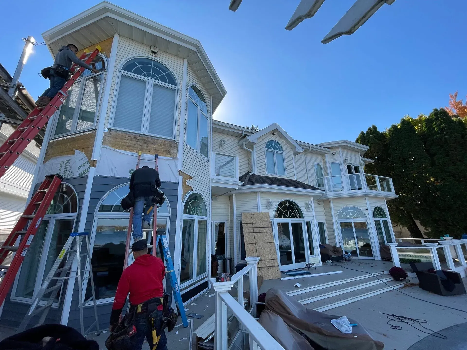 Workers on a ladder repair a large white house with blue trim and arched windows.