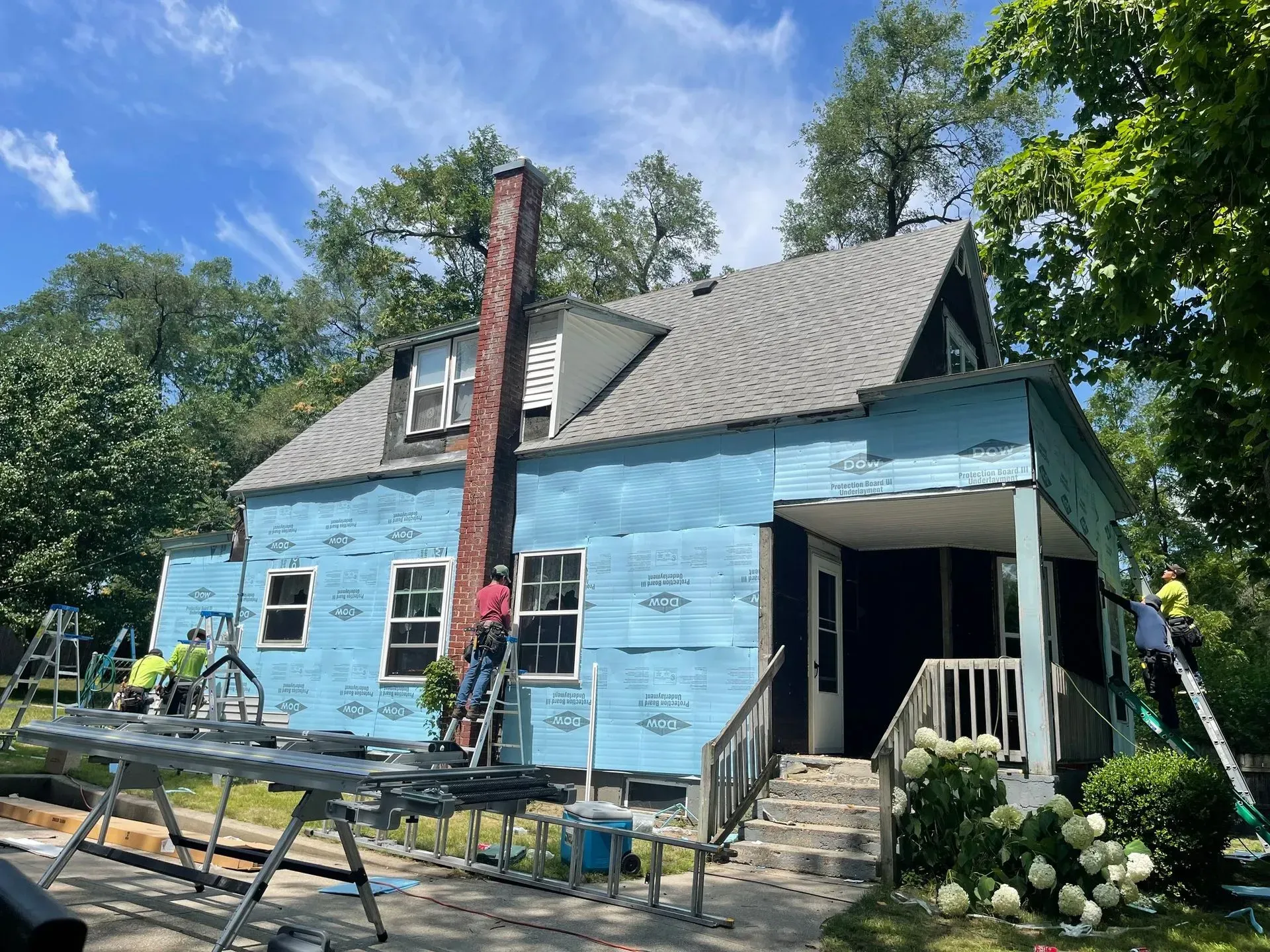 Workers painting a blue house exterior with ladders and scaffolding under a sunny sky