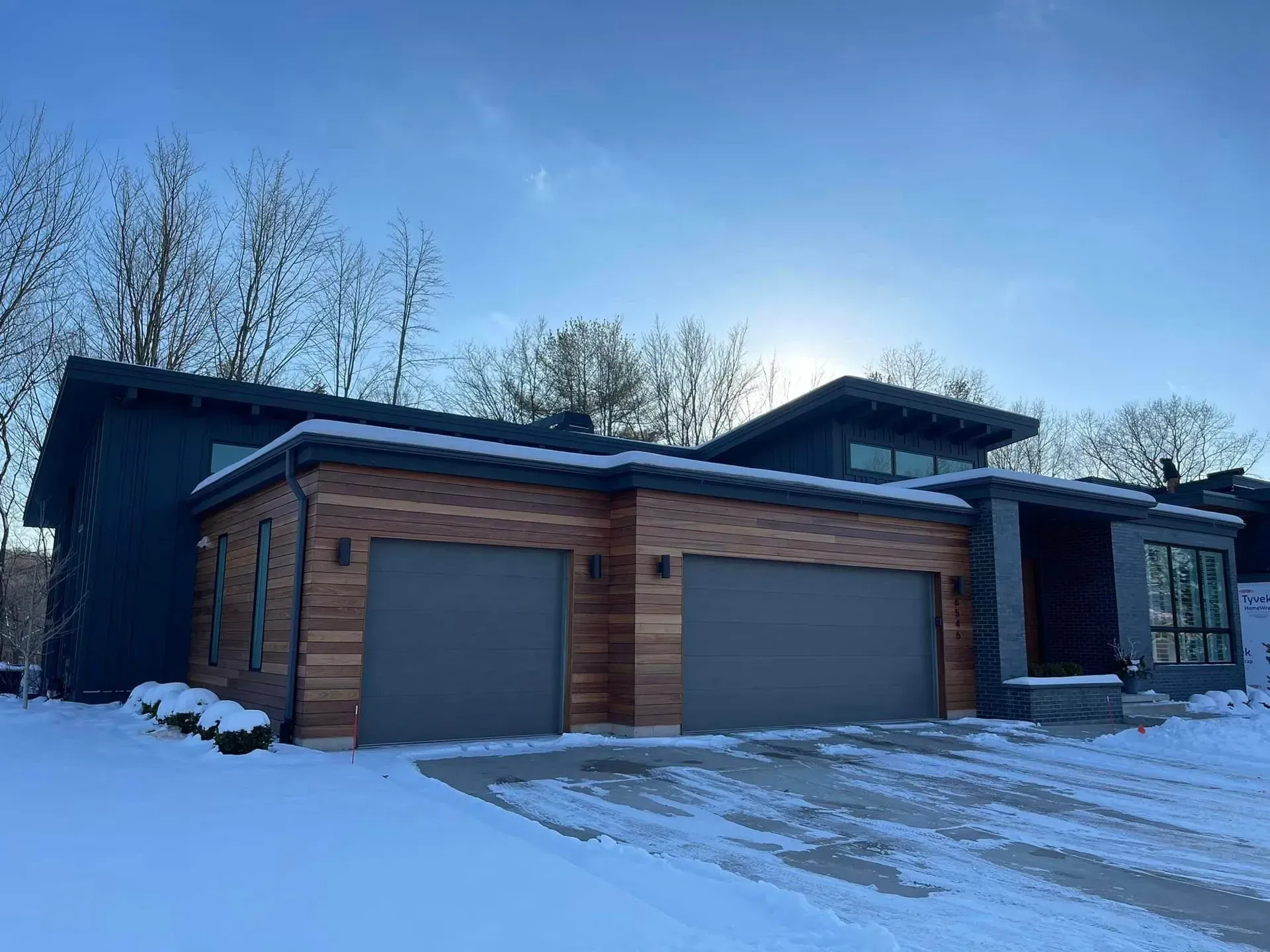 Modern house with wood siding, two garage doors, and a snow-covered driveway under a clear blue sky