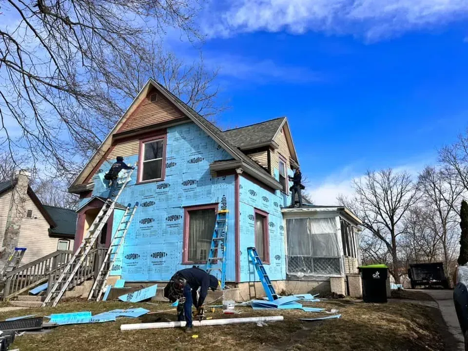Workers wrap a blue house in insulation during exterior renovation.