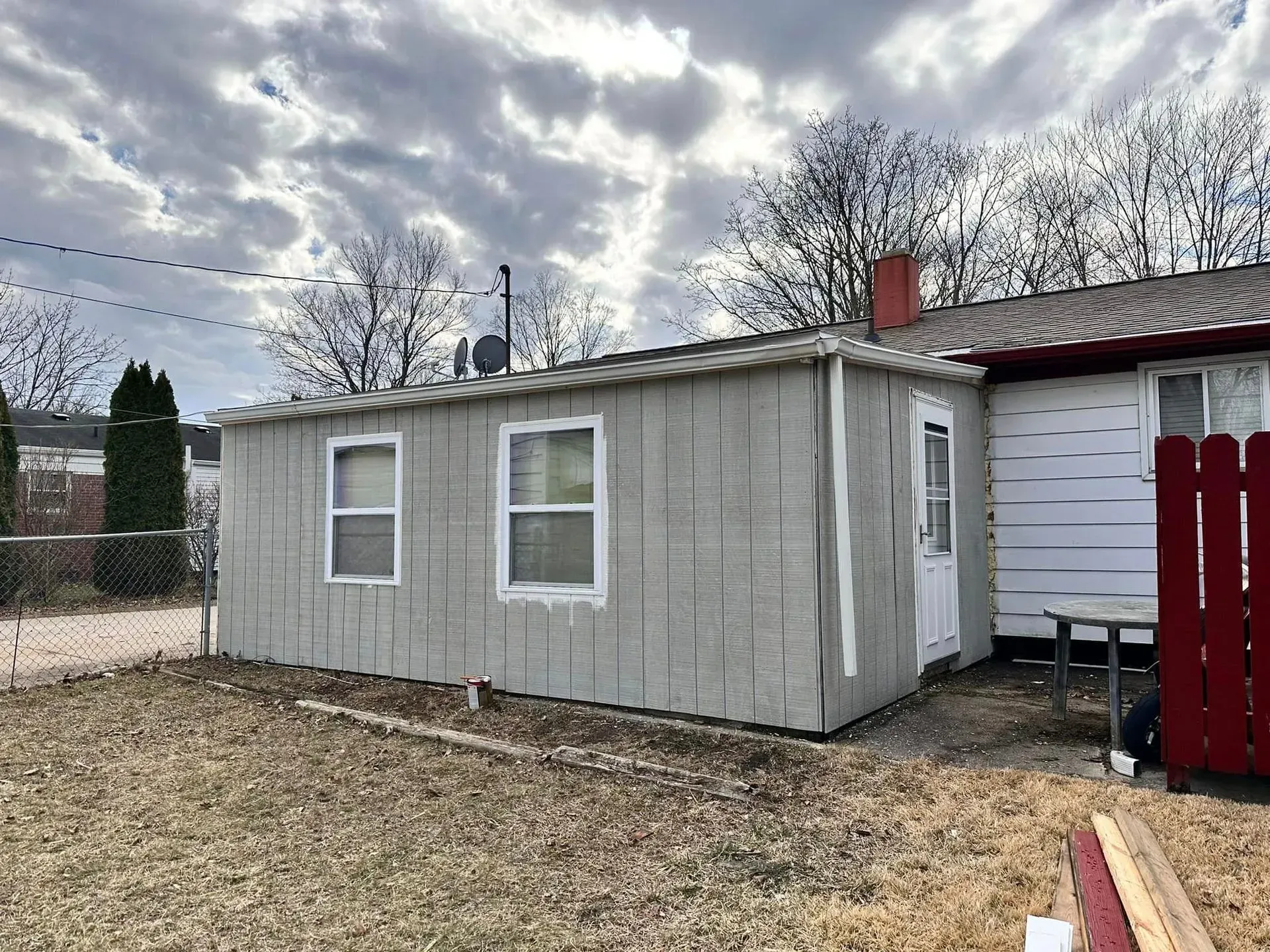 Small gray shed beside a red fence under a cloudy sky, with bare trees in the background.
