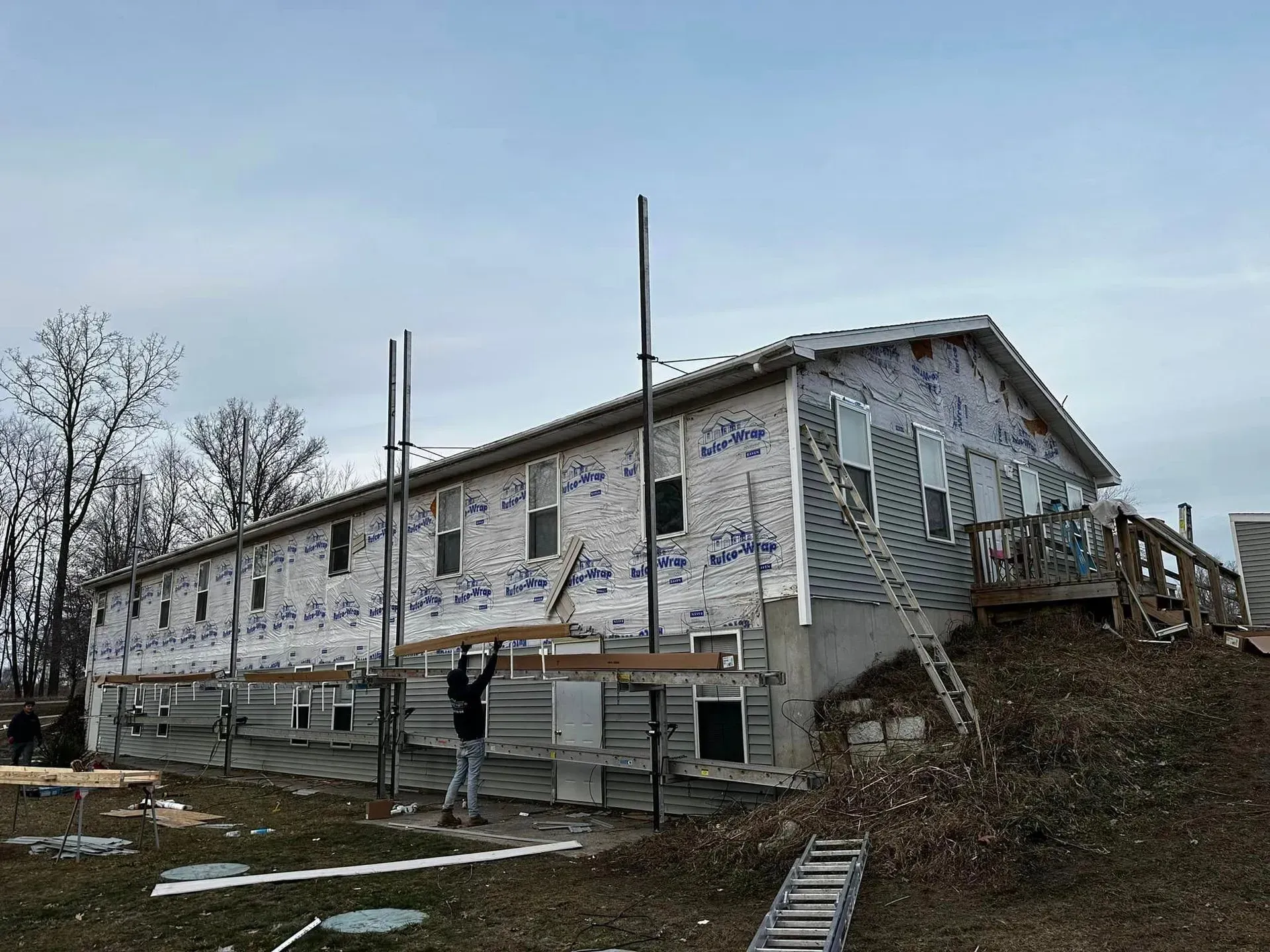 Partially built house wrapped in white housewrap, with scaffolding and workers on a muddy construction site.