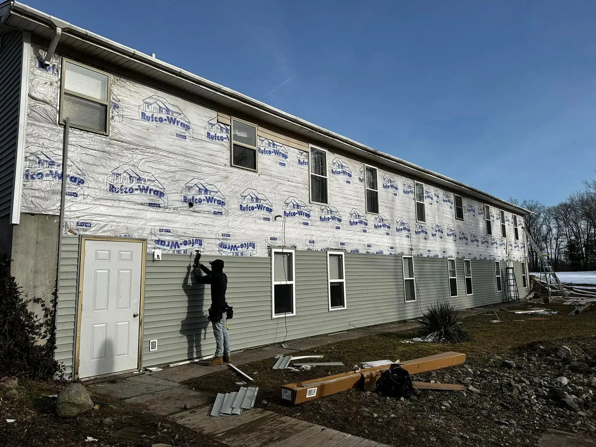 Two-story house with white and green siding, partially wrapped in Tyvek, with debris in the yard.
