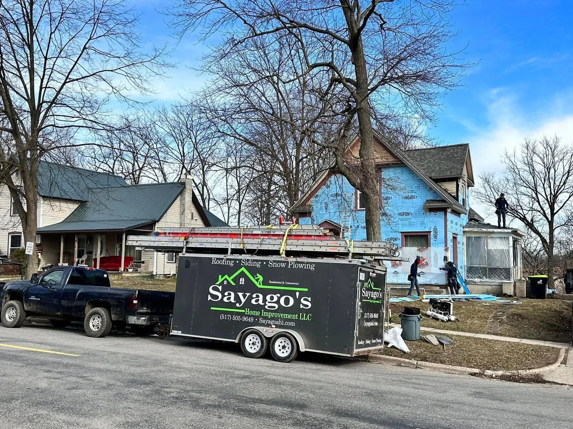 Black Saygo’s trailer parked on a street with a truck, house, and leafless trees in the background.