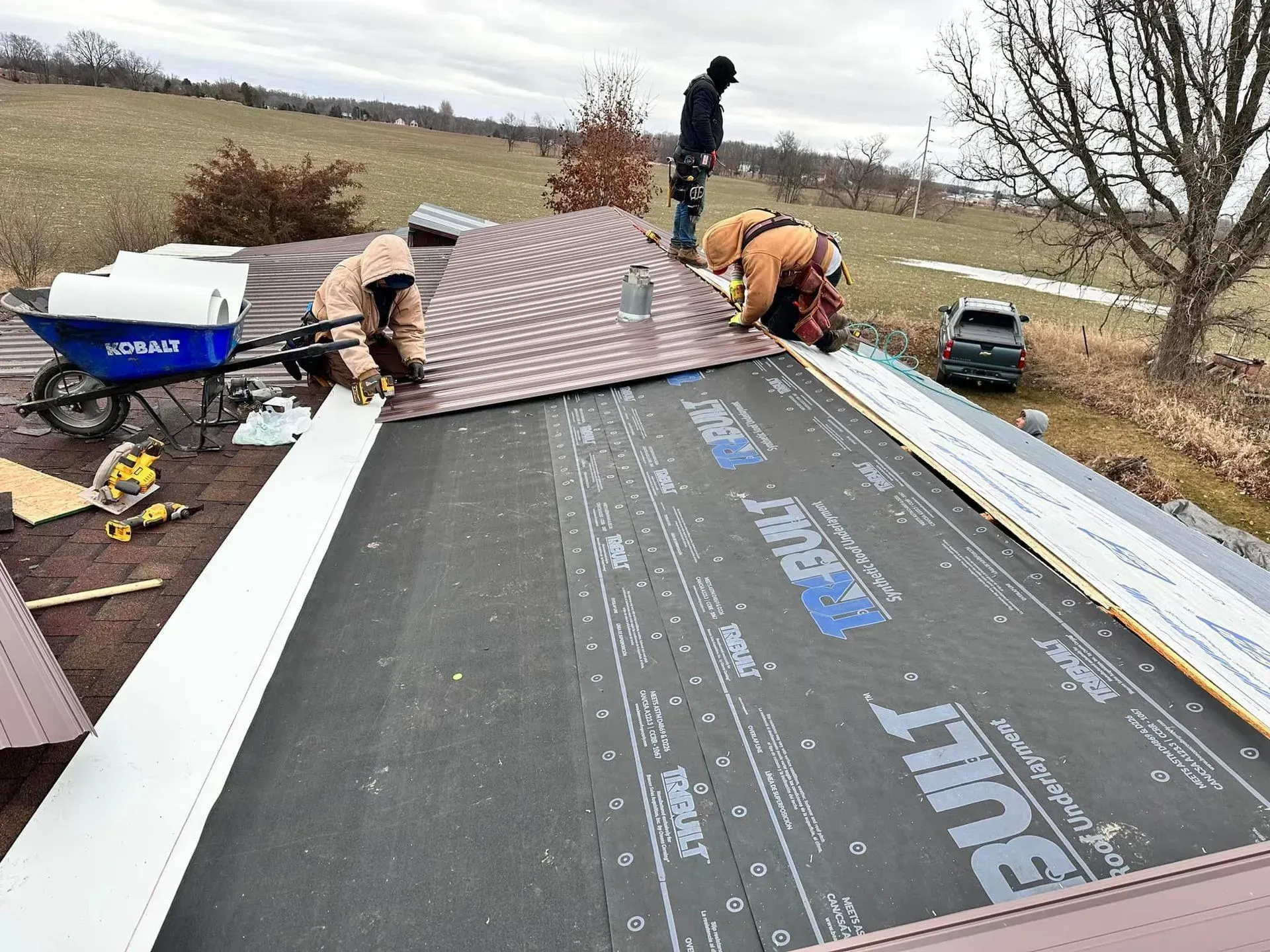 Roofers installing brown shingles on a house roof in a rural area, with underlayment exposed below.