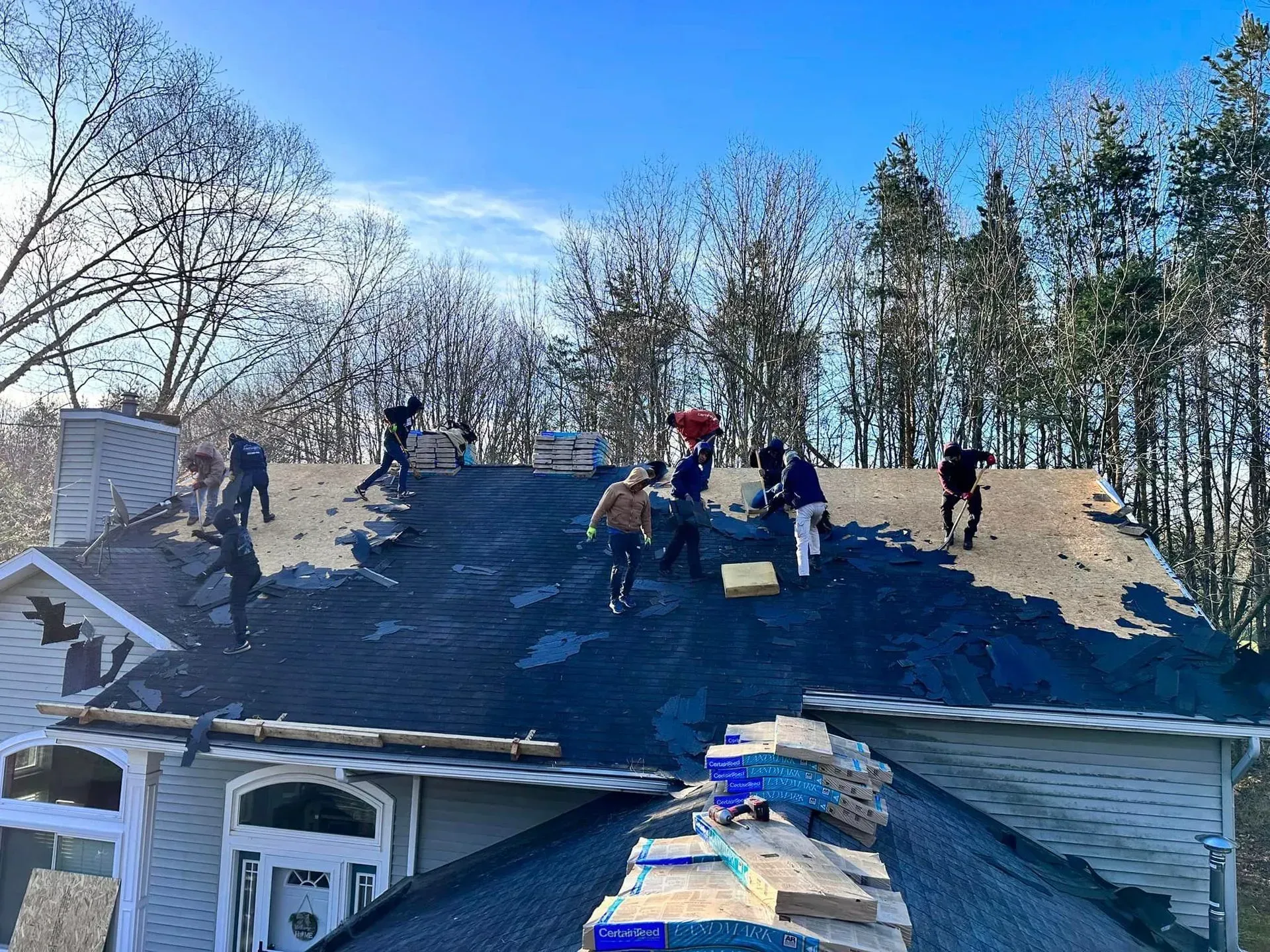 Workers tarping a roof in winter, with several people on the dark blue roof and trees in the background