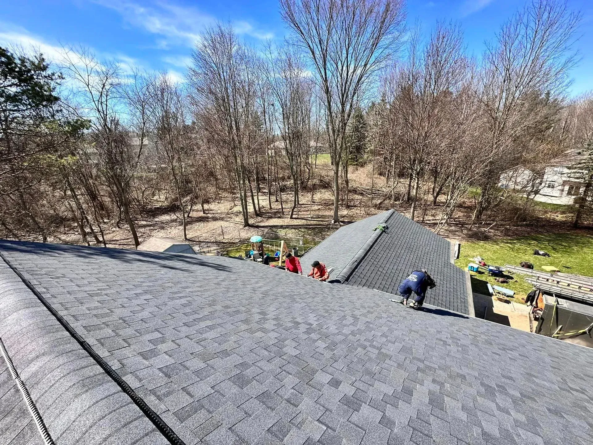 Roofers working on a gray shingled roof beside leafless trees under a blue sky