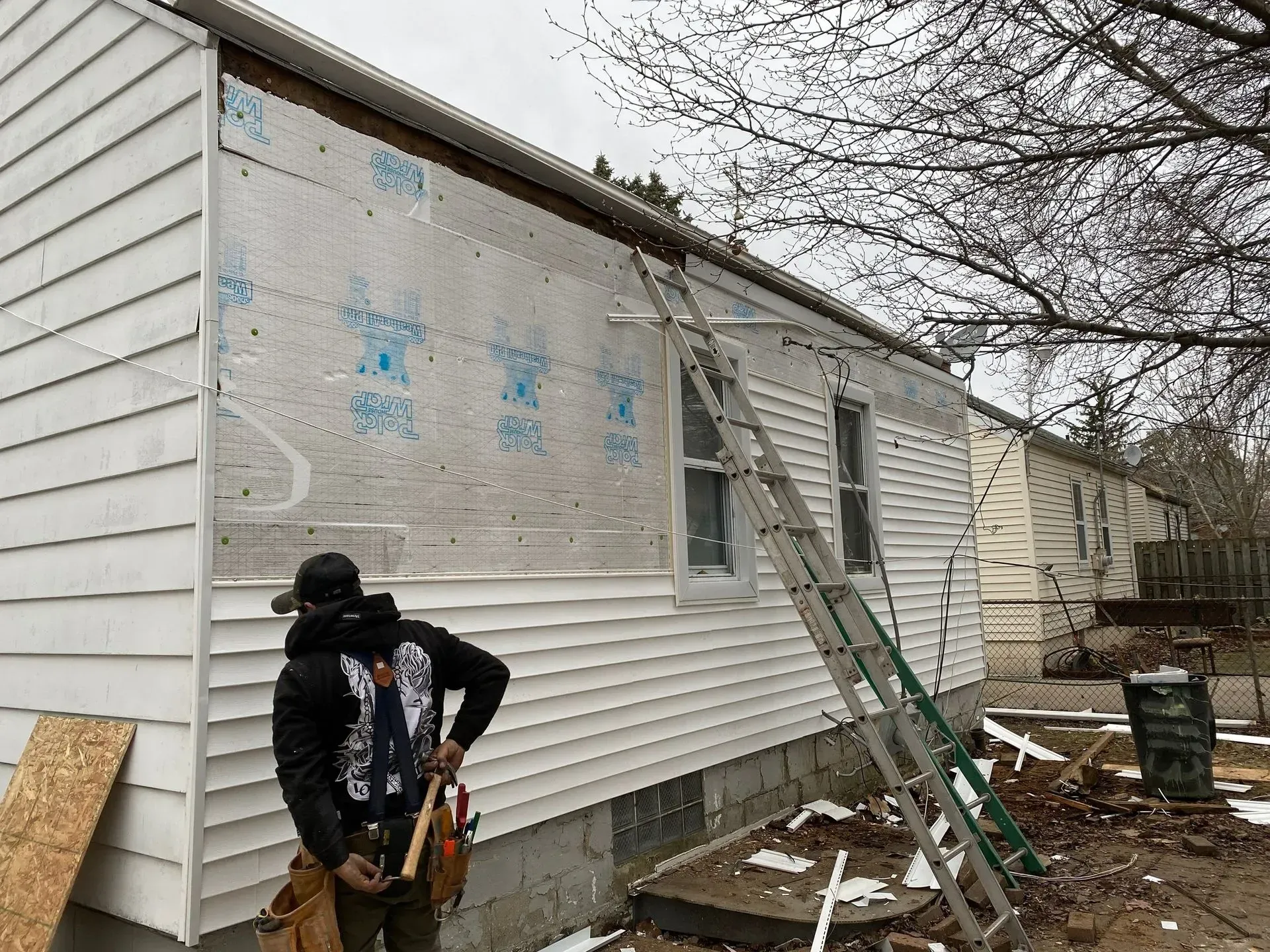 Worker holding a power tool beside a house under repair, with a ladder leaning against the wall.