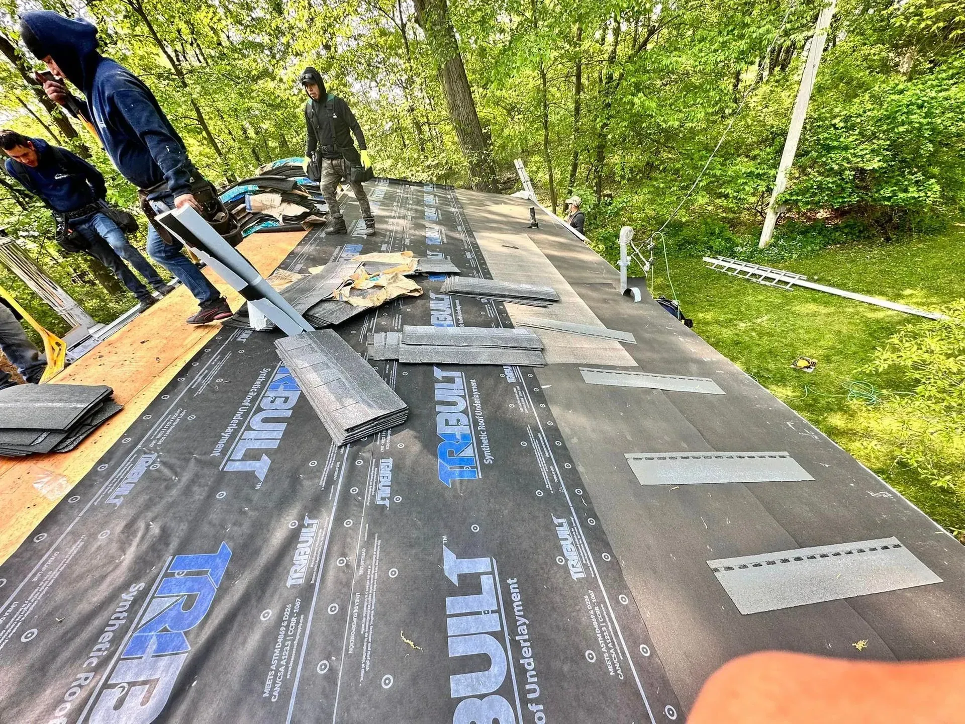 Workers installing black roofing underlayment on a sloped roof in a wooded area