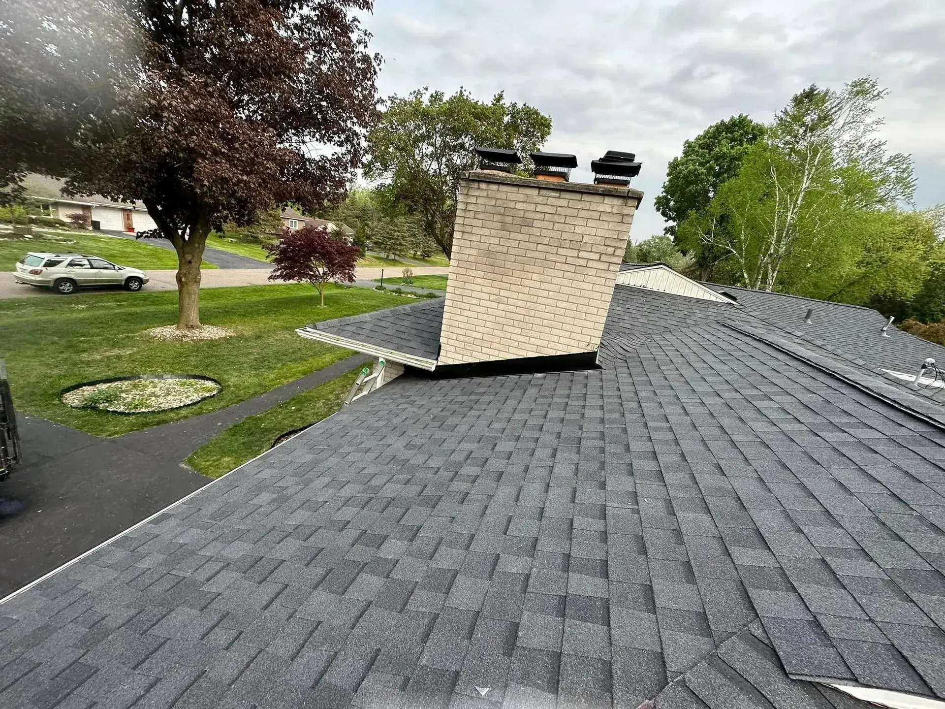 Rooftop with gray shingles and a brick chimney, viewed over a green residential yard with trees.