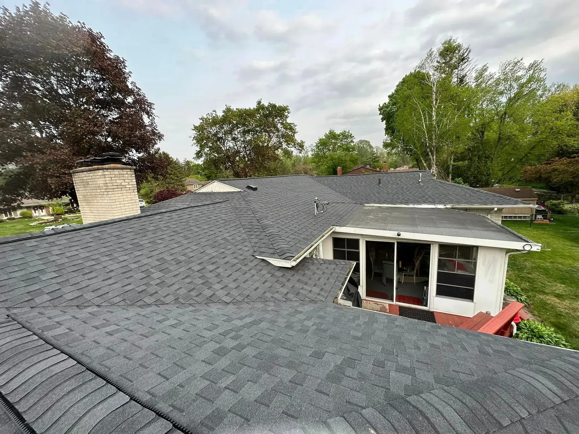Gray shingled roof with damaged section over a house, trees and cloudy sky in the background