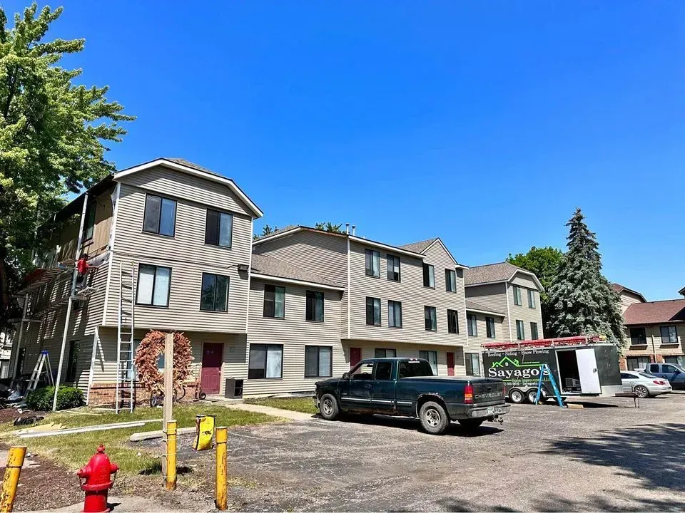 Three-story beige apartment building with parked vehicles, fire hydrant, and trees on a sunny day