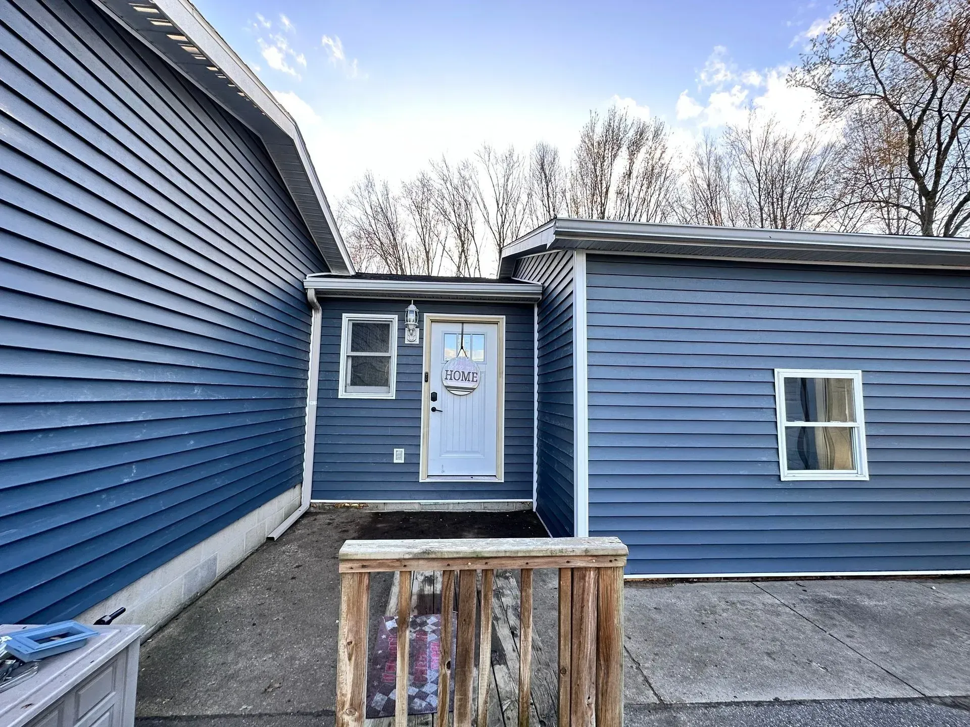 Blue house exterior with a white front door, concrete walkway, and wooden porch railing in the foreground