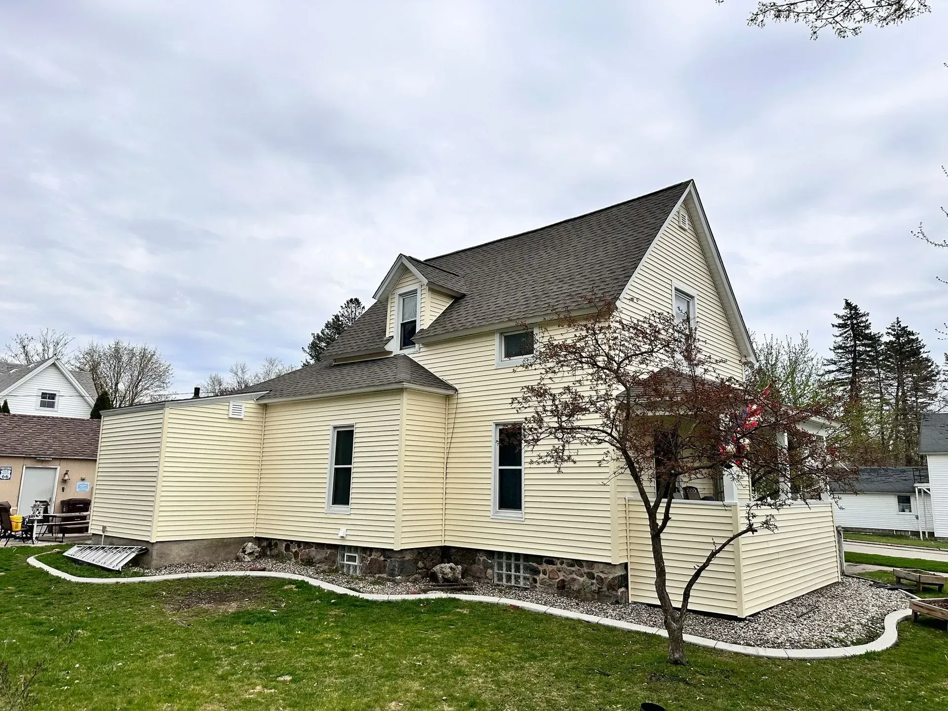 Yellow house with dark roof and curved front addition in a suburban yard under a cloudy sky