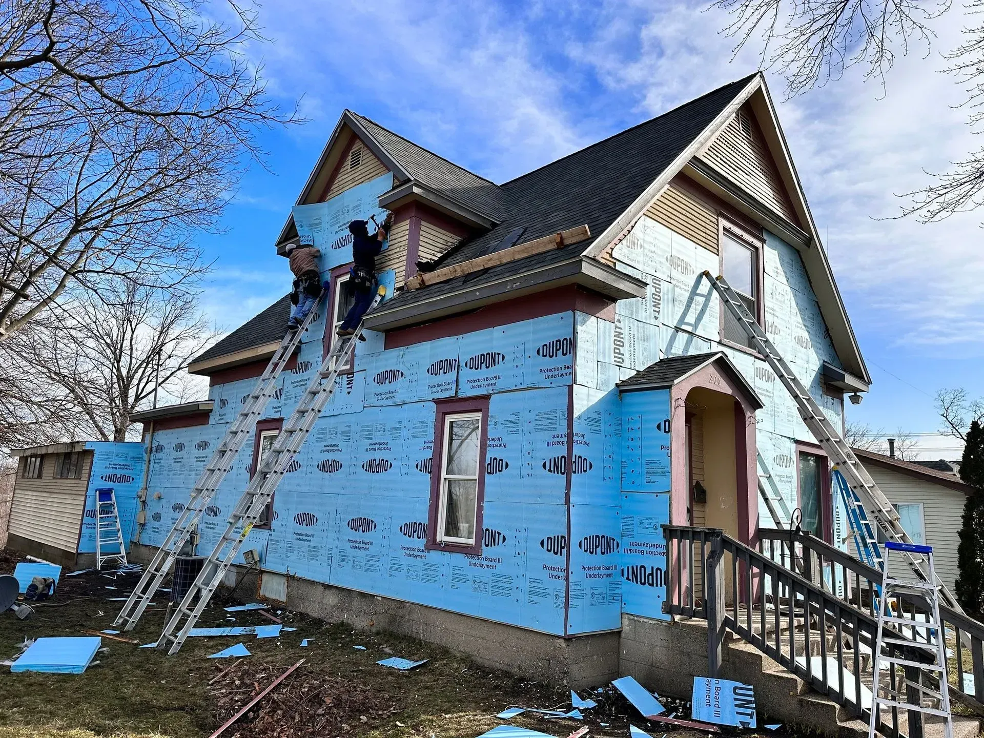 Partially painted house with blue siding, ladder, and workers on the roof under a clear sky