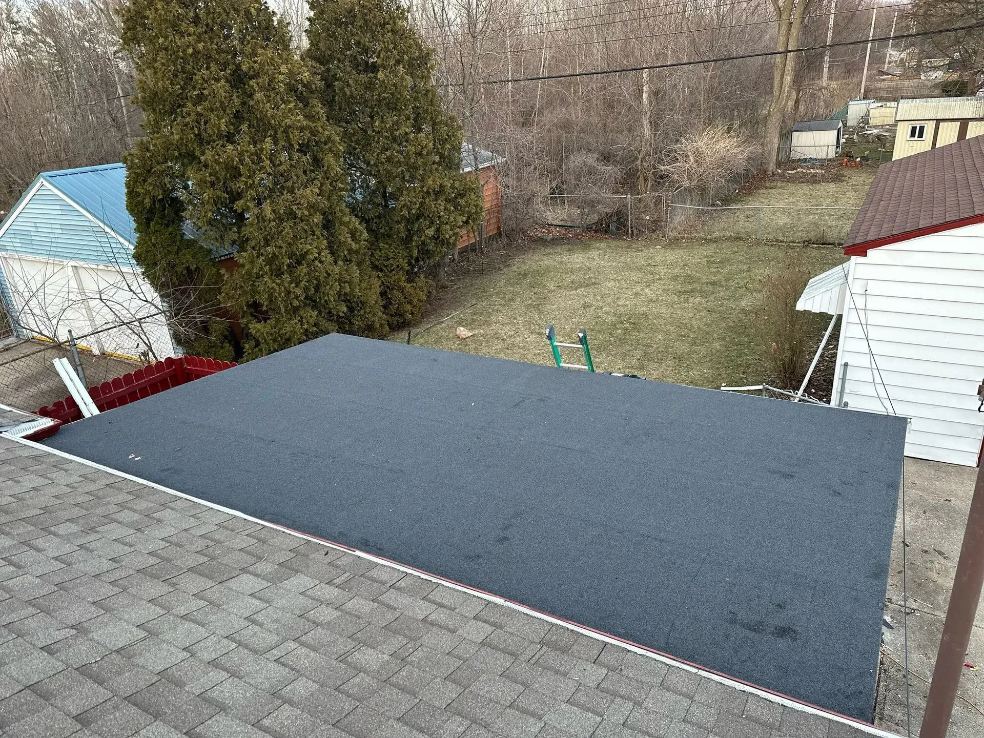 Roof with gray shingles overlooking a yard, with a large dark blue rooftop section and trees in the background