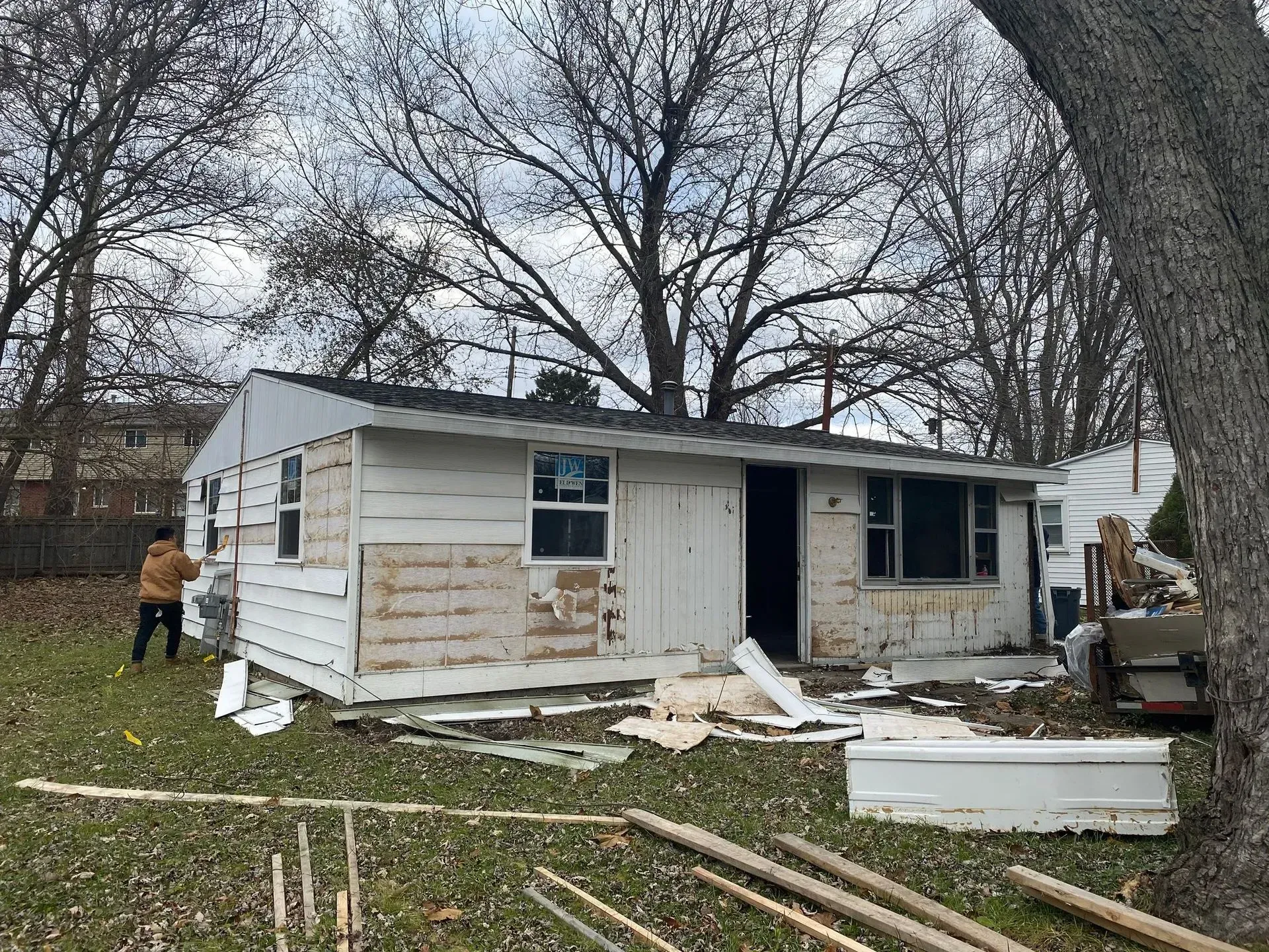 Damaged white house with debris in yard and a person standing to the left under bare trees.