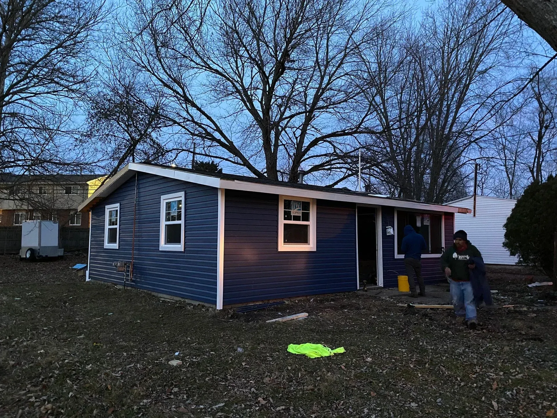Small blue house at dusk with bare trees; a person stands near the porch.