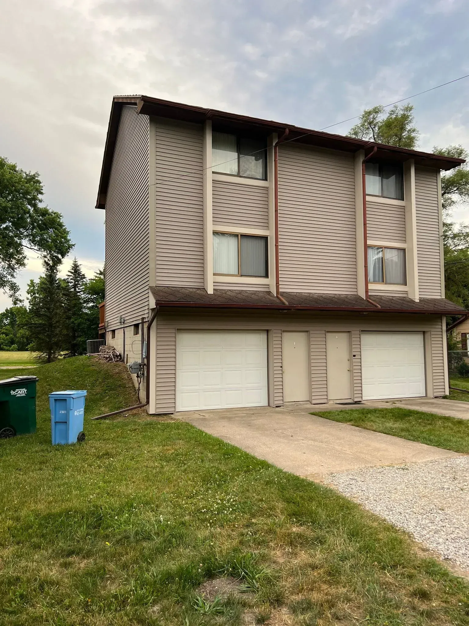Three-story beige townhouse with double garage, gravel driveway, and grassy yard under cloudy sky