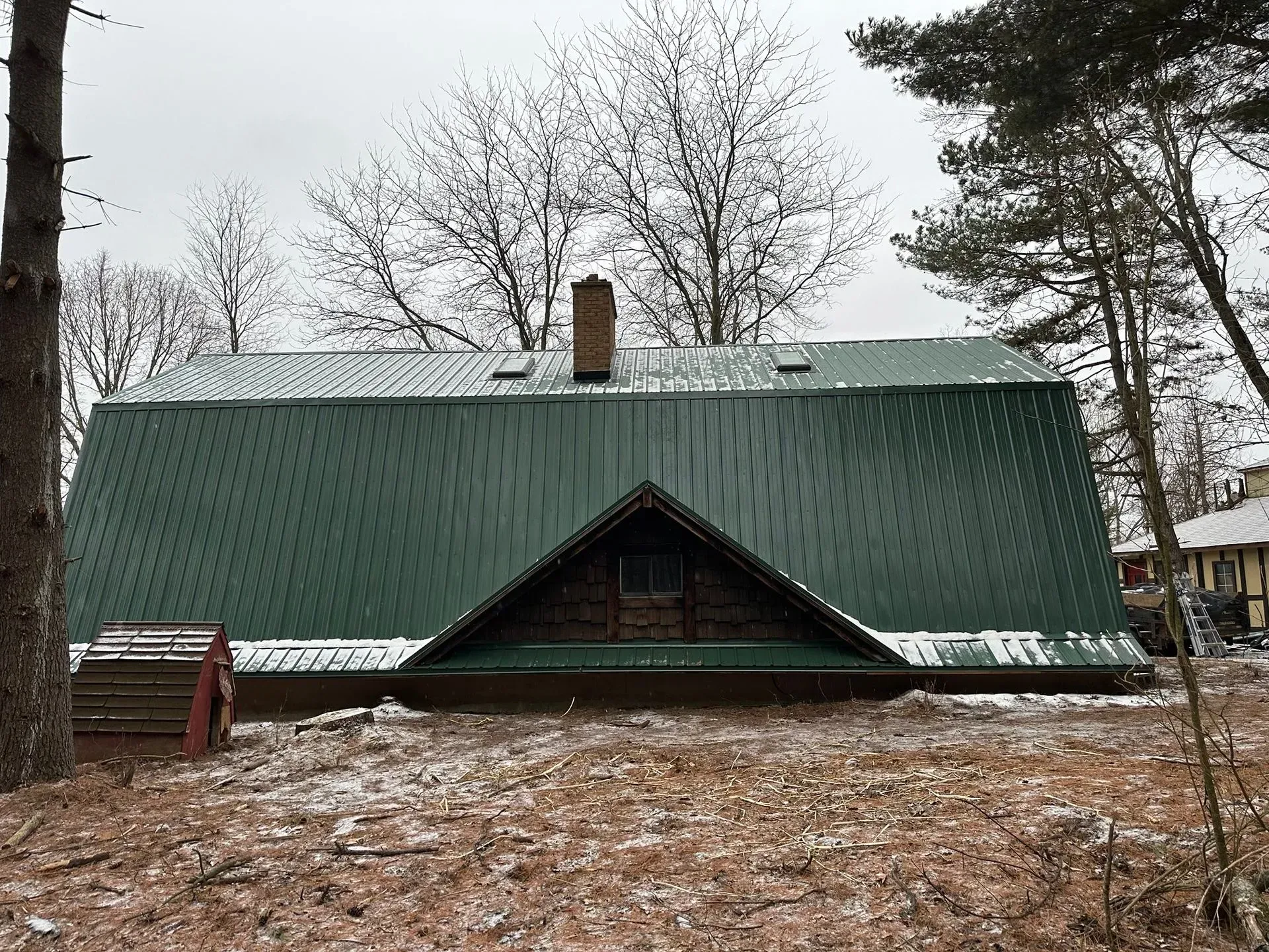 A small green-roofed cabin in a snowy wooded area with leafless trees around it.