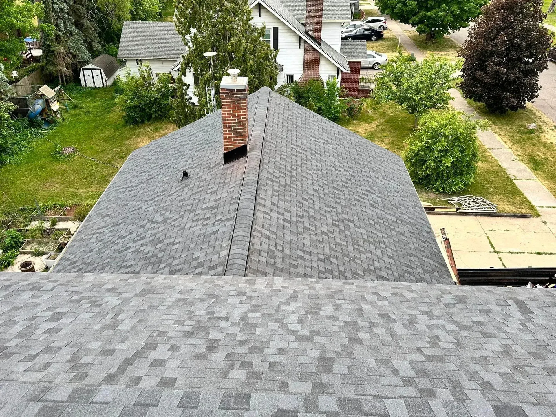 View from a roof overlooking a gray shingled house, chimney, and leafy backyard with paths and neighboring homes.