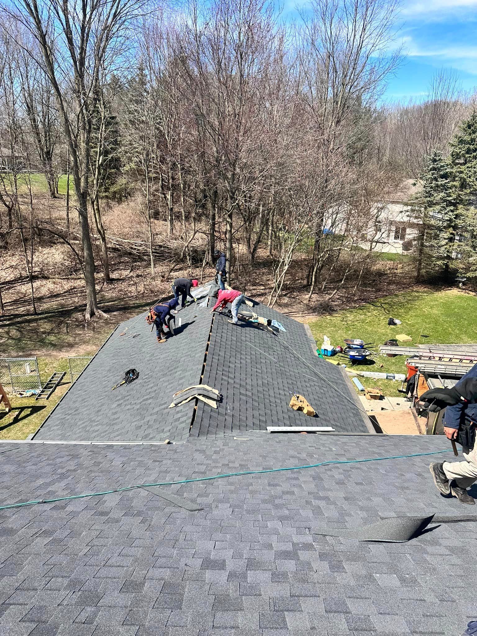 Workers installing black shingles on a steep roof amid trees on a sunny day
