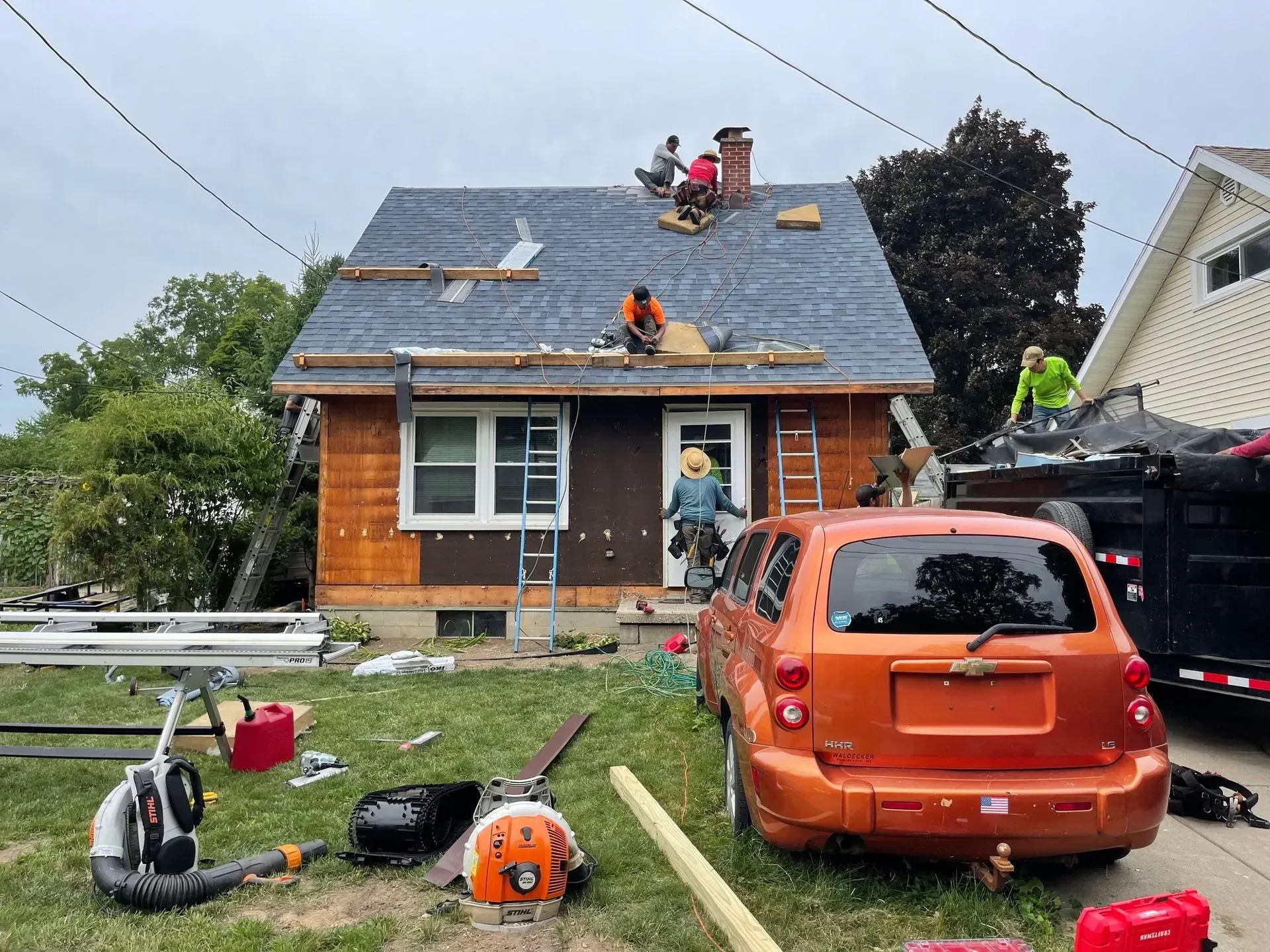 Workers repairing a house roof with tools and debris in the yard, orange car parked in front