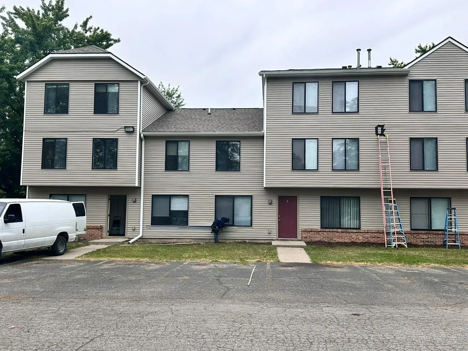 Three-story apartment building with gray siding, a white van parked outside, and a ladder leaning against the right side.