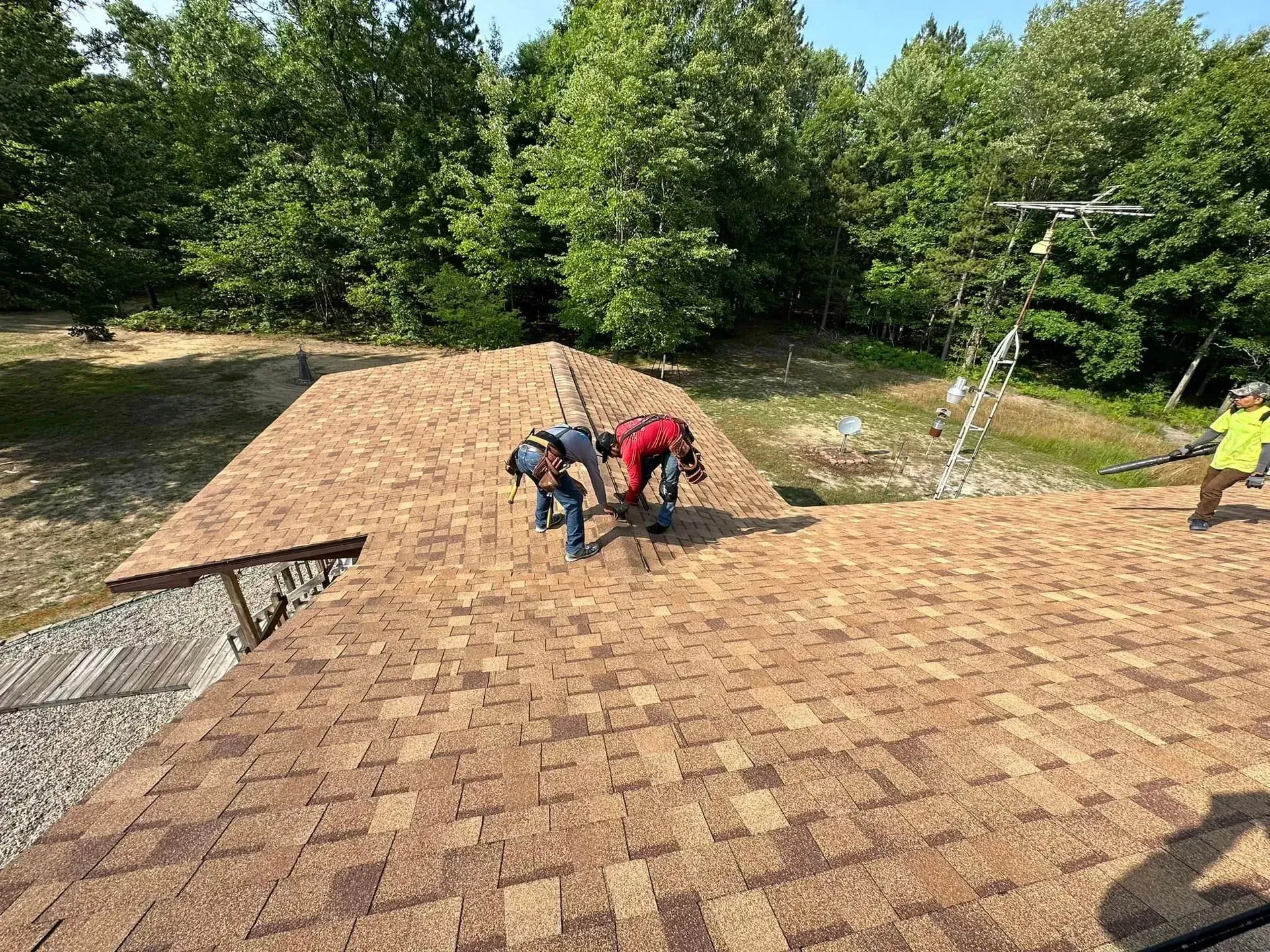 Workers installing shingles on a brown roof with a wooded yard and sprinklers in the background