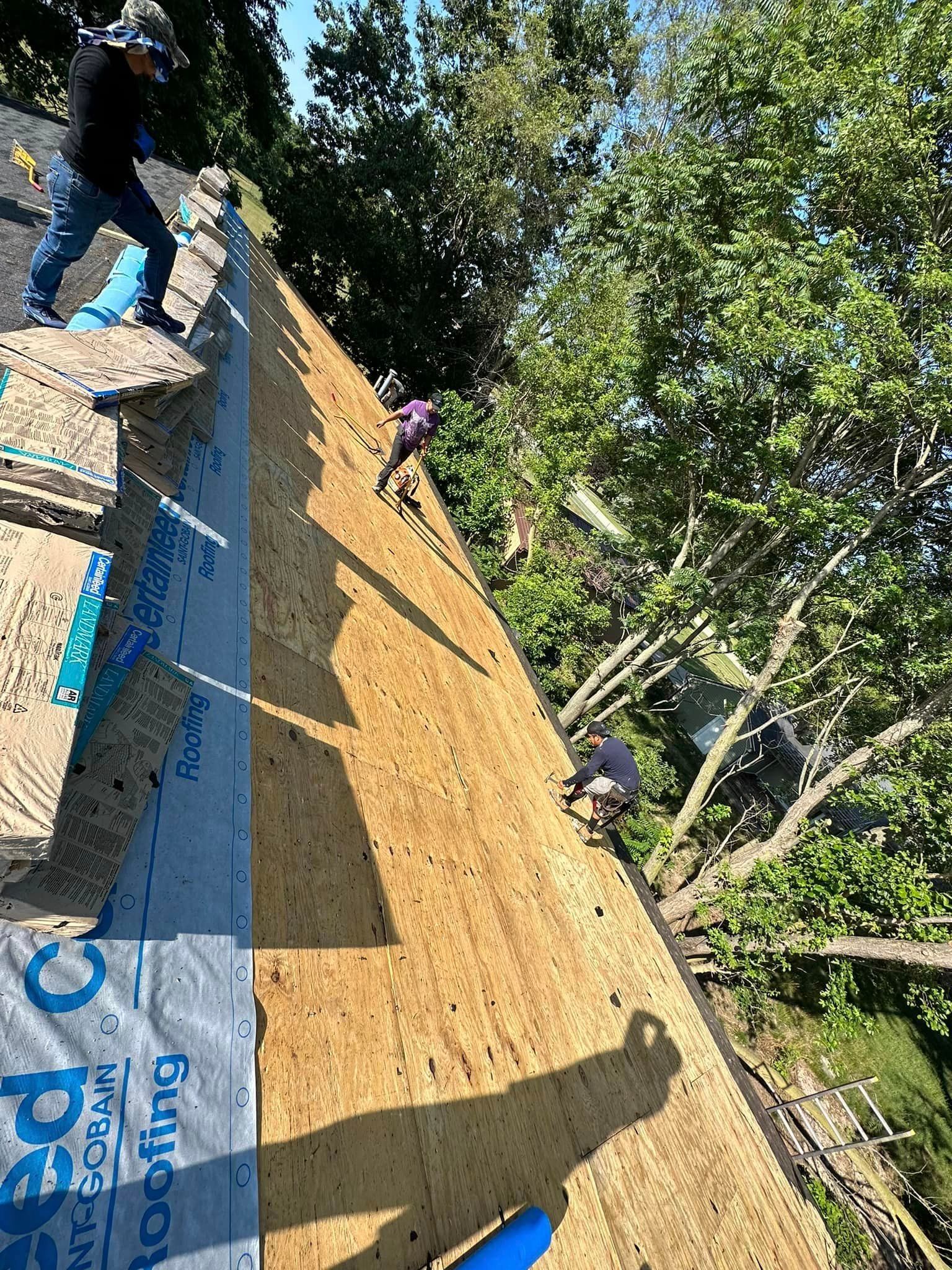 Workers on a sloped rooftop under construction, with blue tarp and trees nearby.