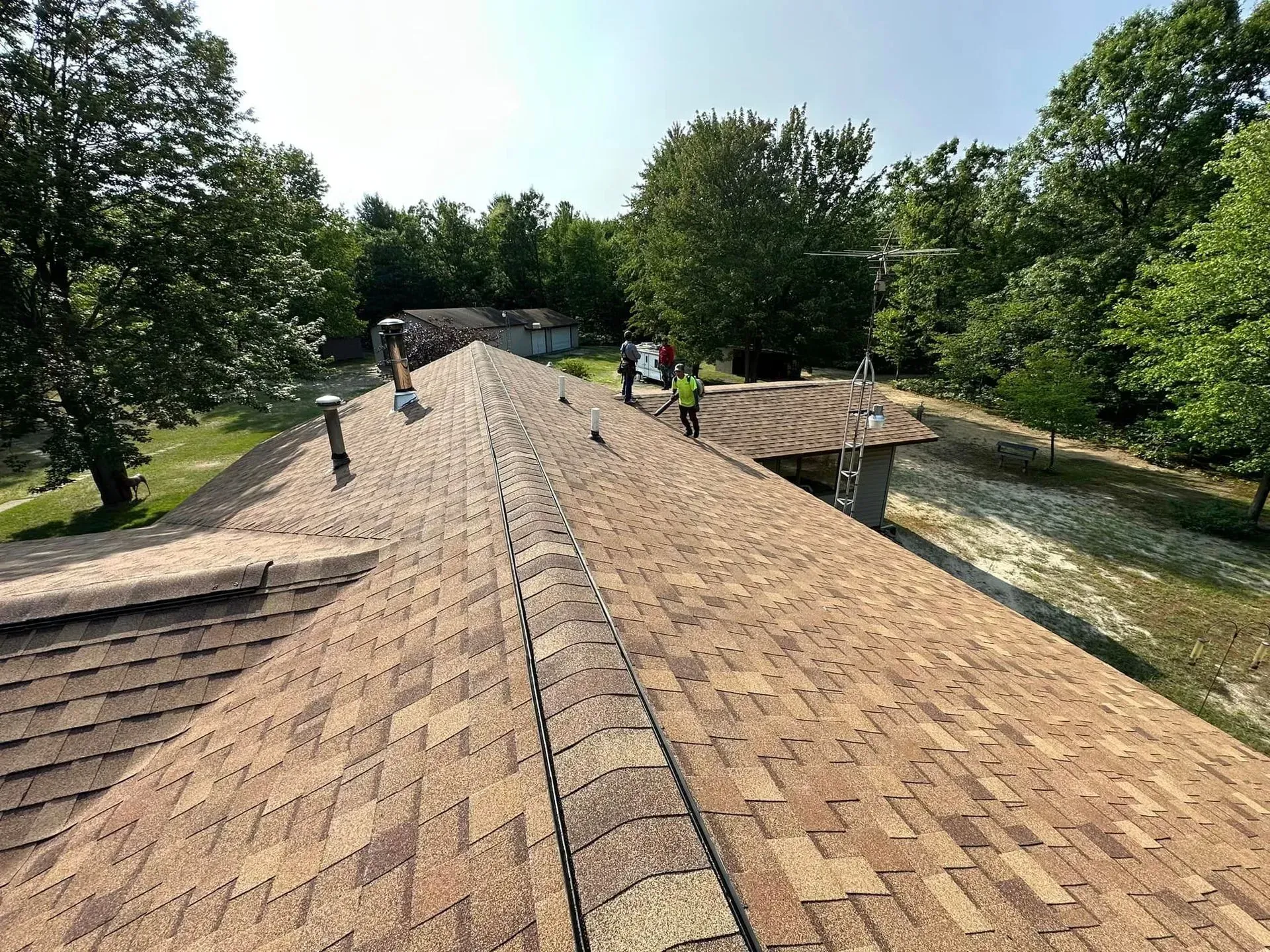 Workers repairing a shingled roof in a wooded area on a sunny day